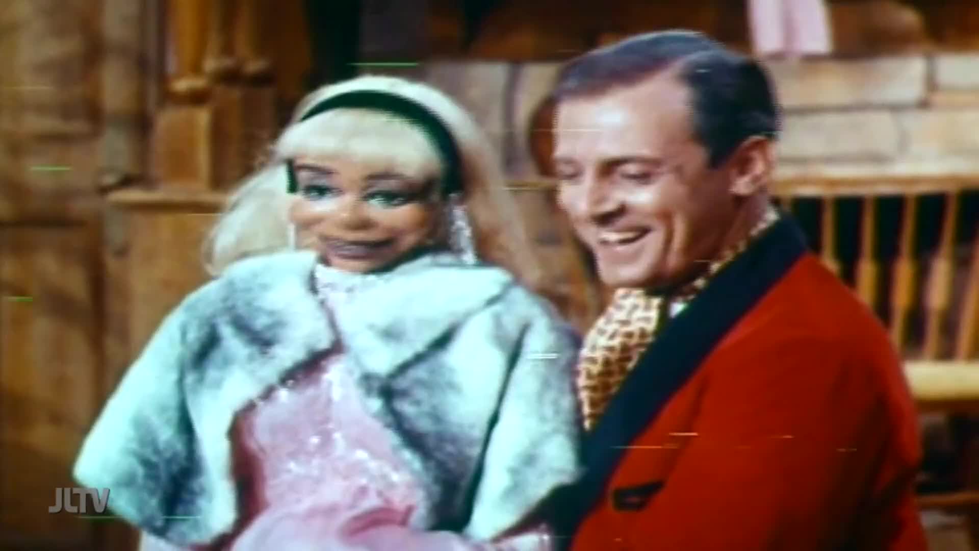 A man in a red jacket smiles beside a doll wearing a fur stole. The scene appears to be from a program on Jewish Life Television.
