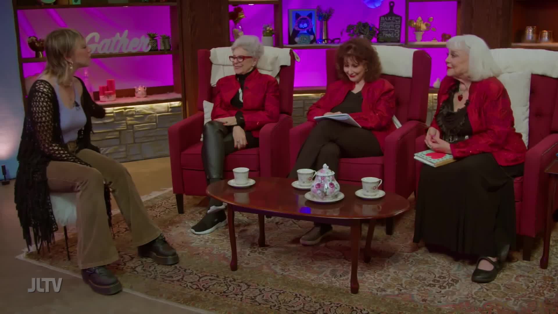 On a set for Jewish Life Television, four women sit in armchairs, chatting around a small table set with tea cups. The woman on the left gestures as she speaks, while the others listen, some holding notes or a book.
