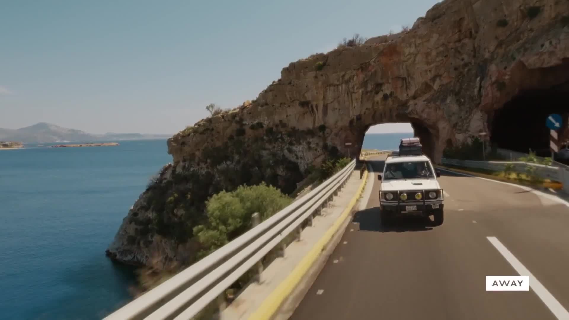 A white SUV, loaded with luggage, drives toward a tunnel carved through a rocky cliffside. The road hugs the coast, with the blue sea visible on the left.
