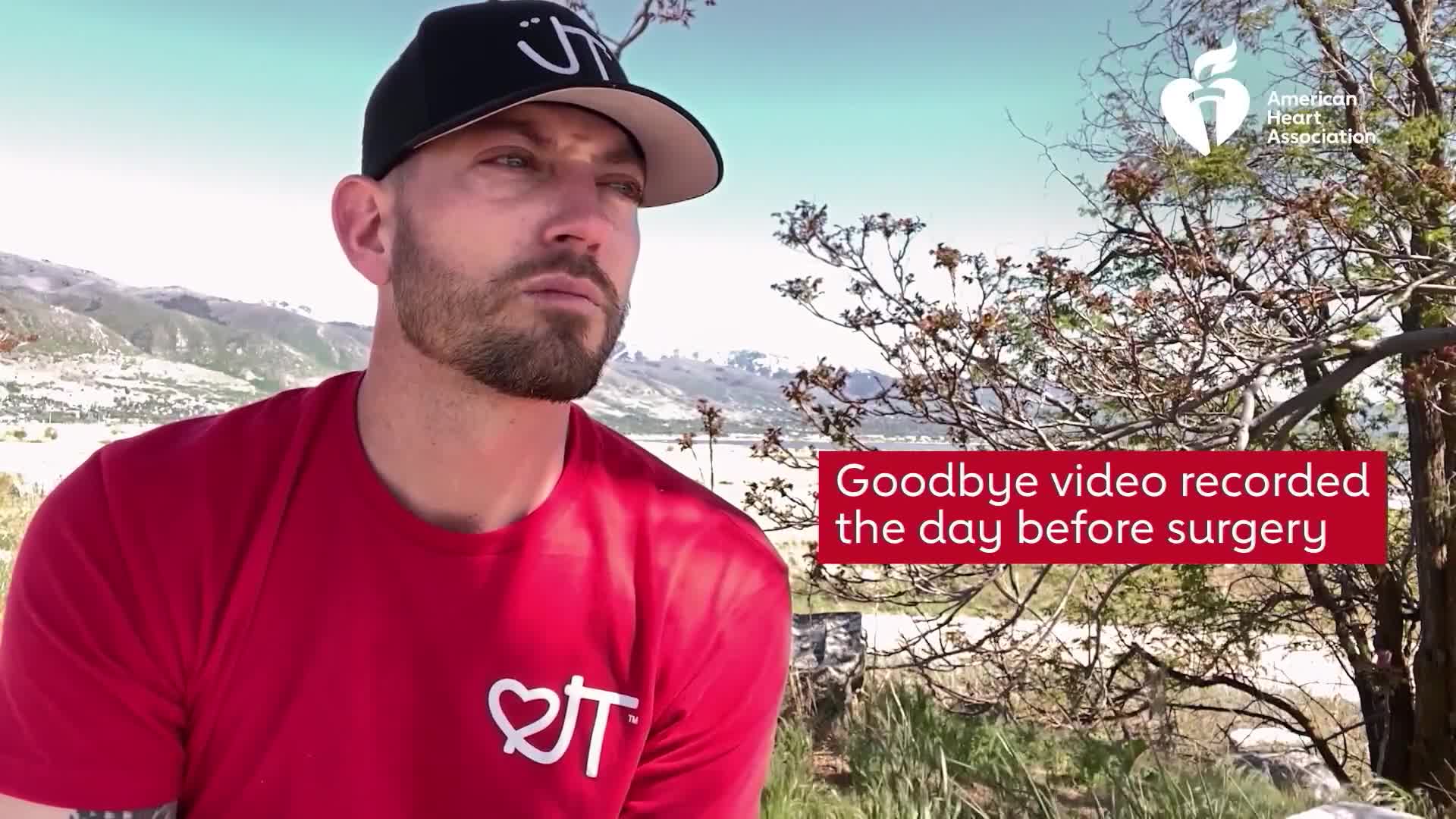 A man in a red shirt and a black cap looks off-camera, the text "Goodbye video recorded the day before surgery" appears on the screen. The scene is set outdoors, with mountains and trees in the background.

