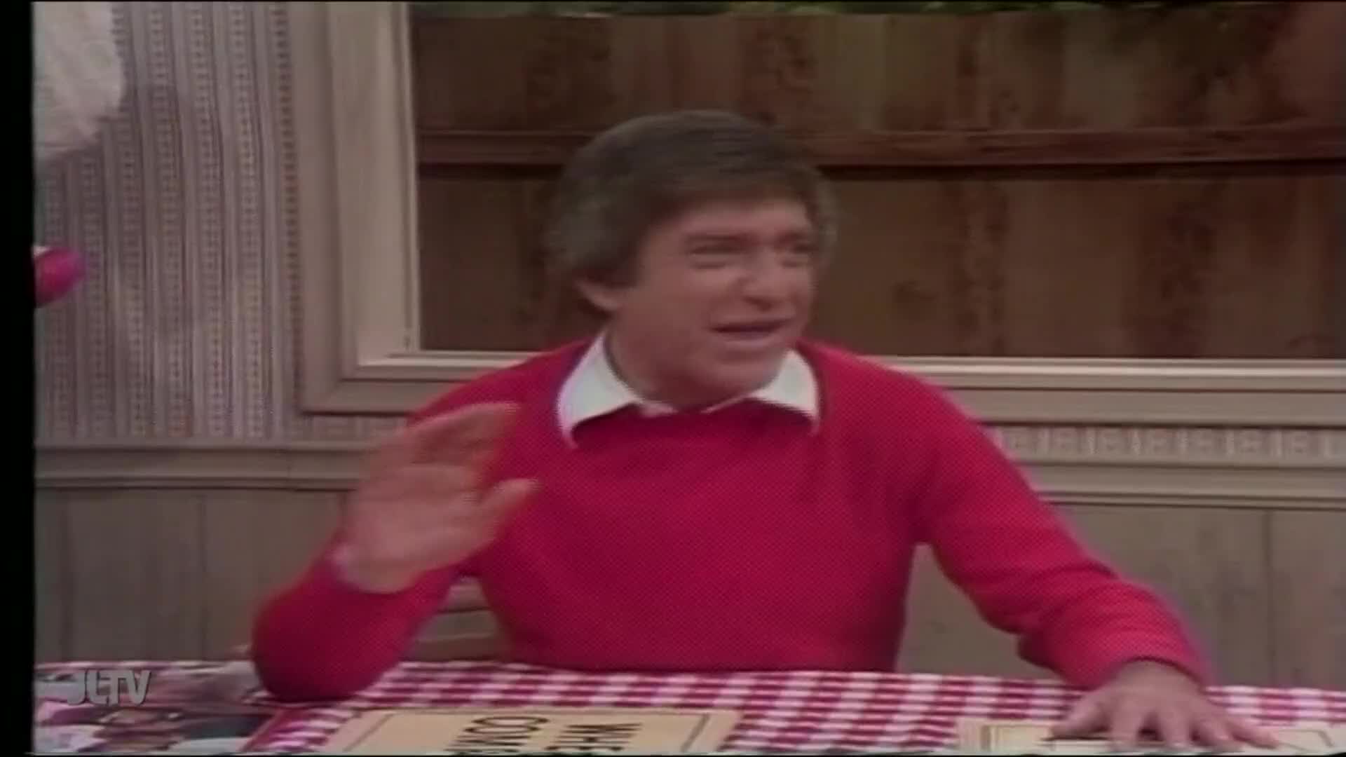 A man in a red sweater waves his hand, seated at a table with a red-and-white checkered tablecloth. The JTLV logo is visible in the corner, suggesting this is a program on Jewish Life Television in the United States.
