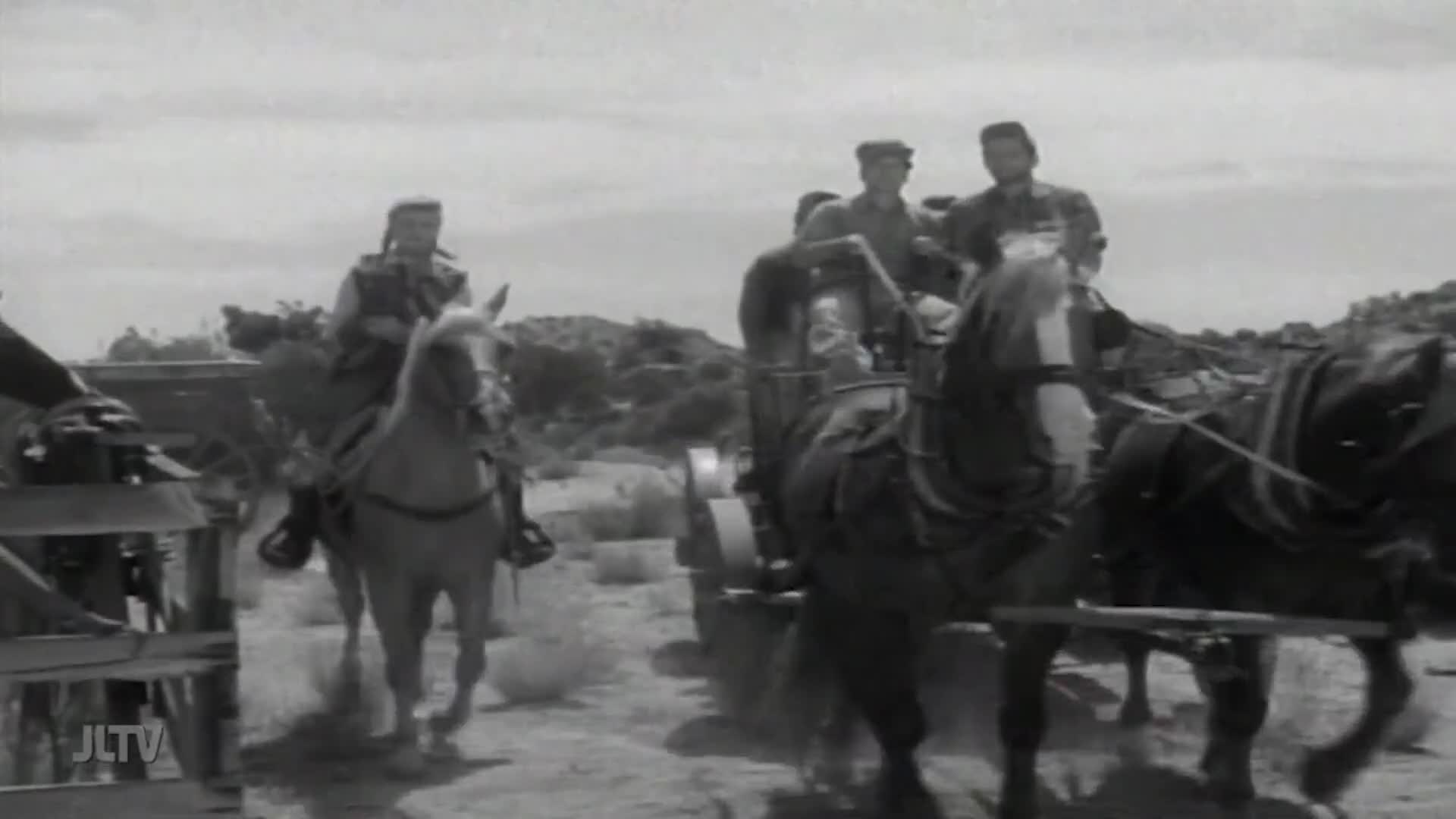 A horse-drawn wagon carrying several men moves across a dusty landscape. A rider on a separate horse, also heading in the same direction, follows alongside.
