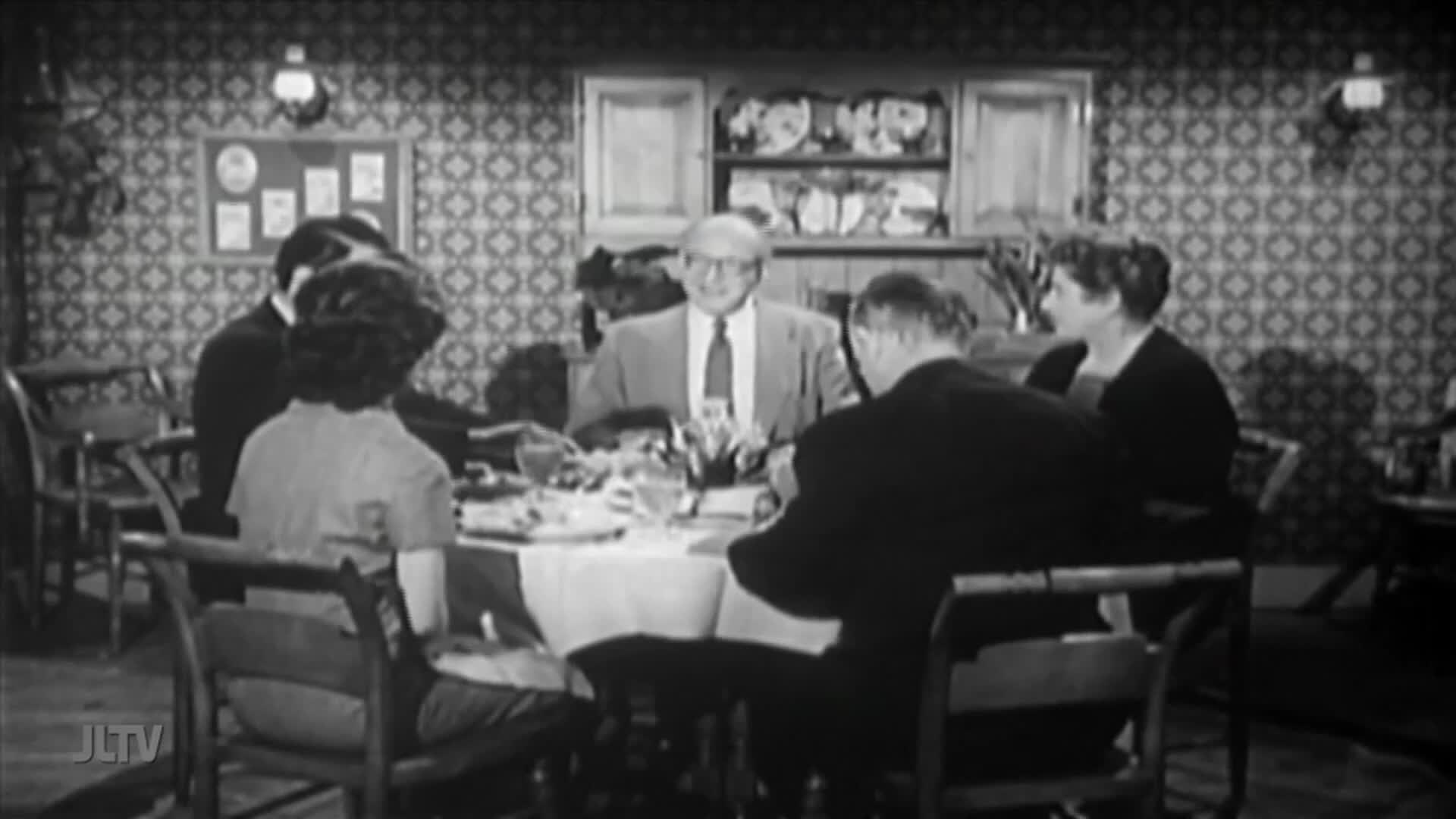 A group of people are gathered around a table, the host in a suit and tie. They're seated in a room with patterned wallpaper, likely a scene from a broadcast on Jewish Life Television in the United States.
