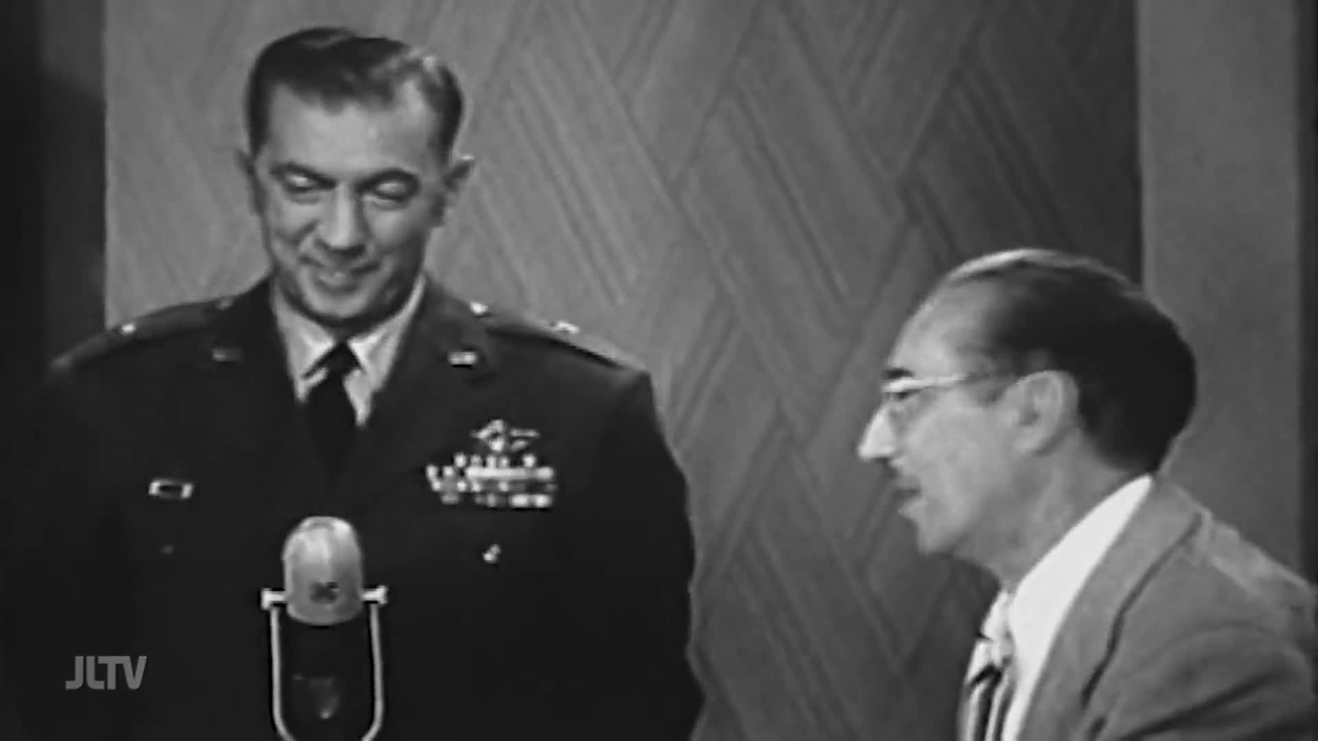 A man in a military uniform smiles, standing near a microphone on a set for Jewish Life Television. Beside him, another man in a suit listens intently.
