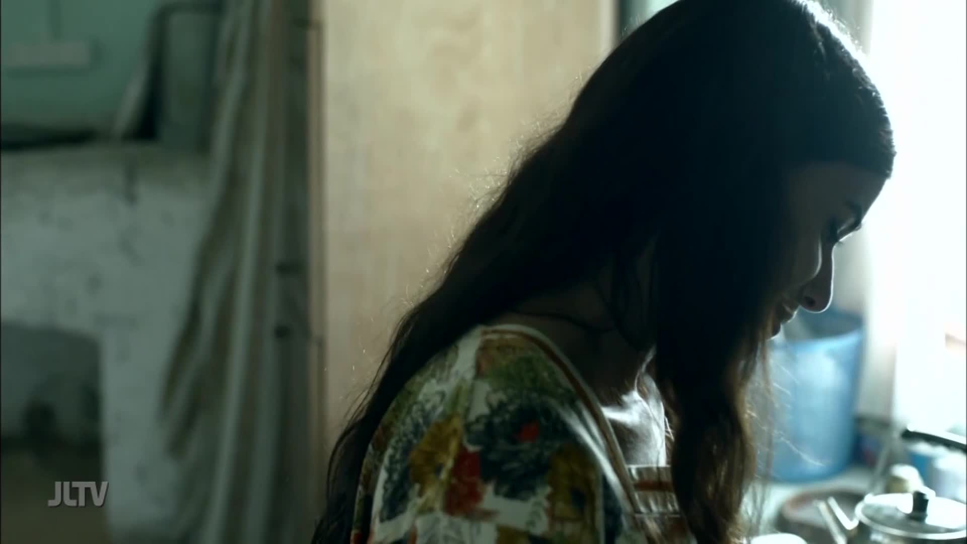 A woman with long, dark hair is looking down, her face lit by the light. She's wearing a floral-patterned top, and the logo of JLTV is visible in the corner.
