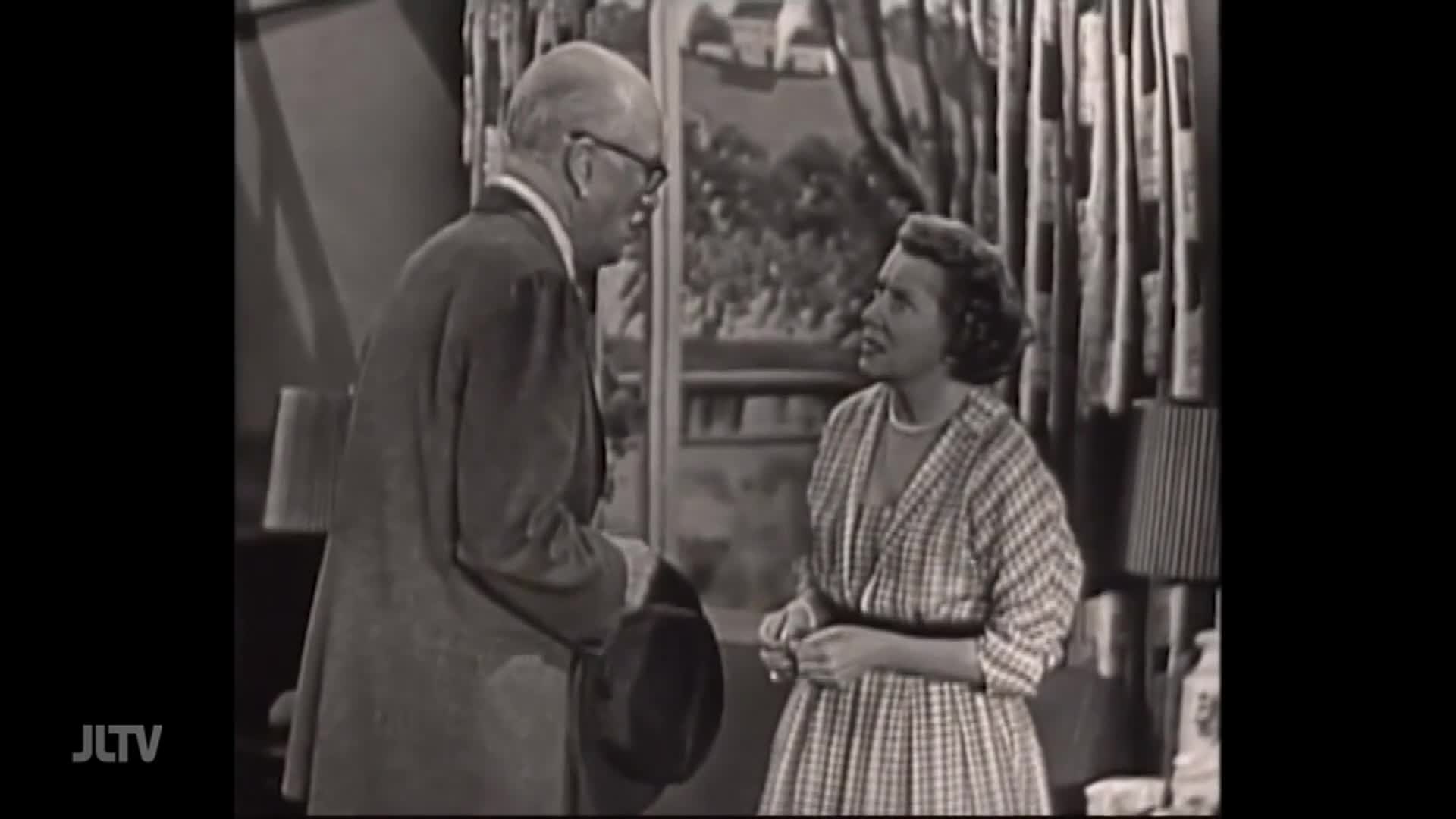A man in a suit and glasses holds a hat, speaking to a woman in a patterned dress. She listens intently, her brow furrowed as if she's concerned or confused.
