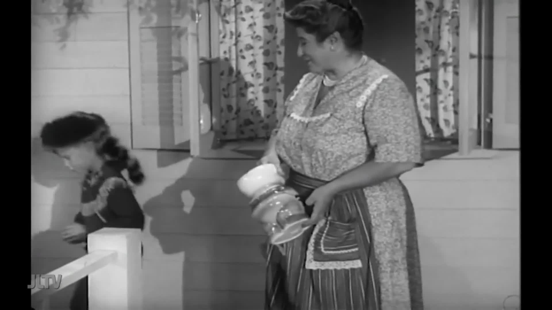 A woman in a floral dress carries a large glass container, her apron pocket visible. A young girl, wearing a dark hat and coat, stands by a white railing on the porch.
