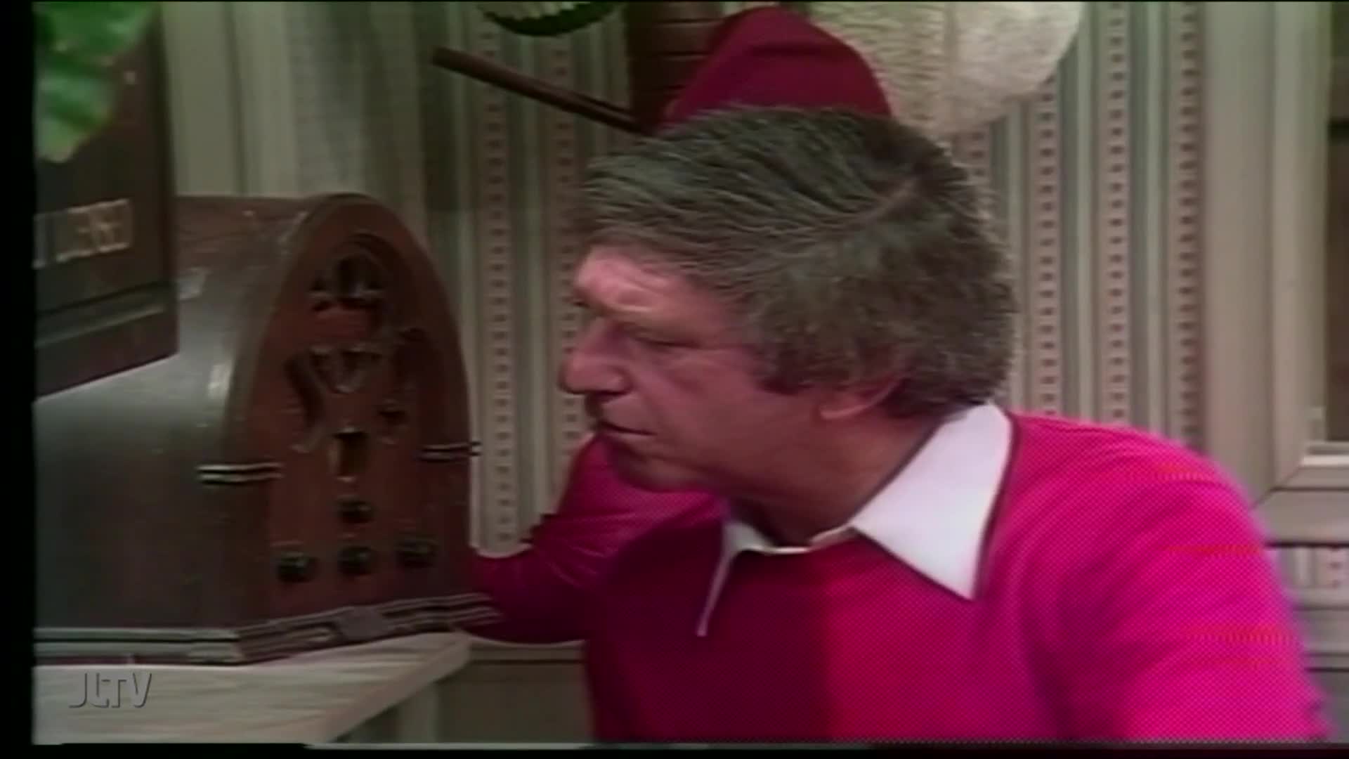 A man in a pink sweater is intently focused on an old wooden radio. He seems to be adjusting the knobs, perhaps hoping to catch a broadcast on Jewish Life Television.
