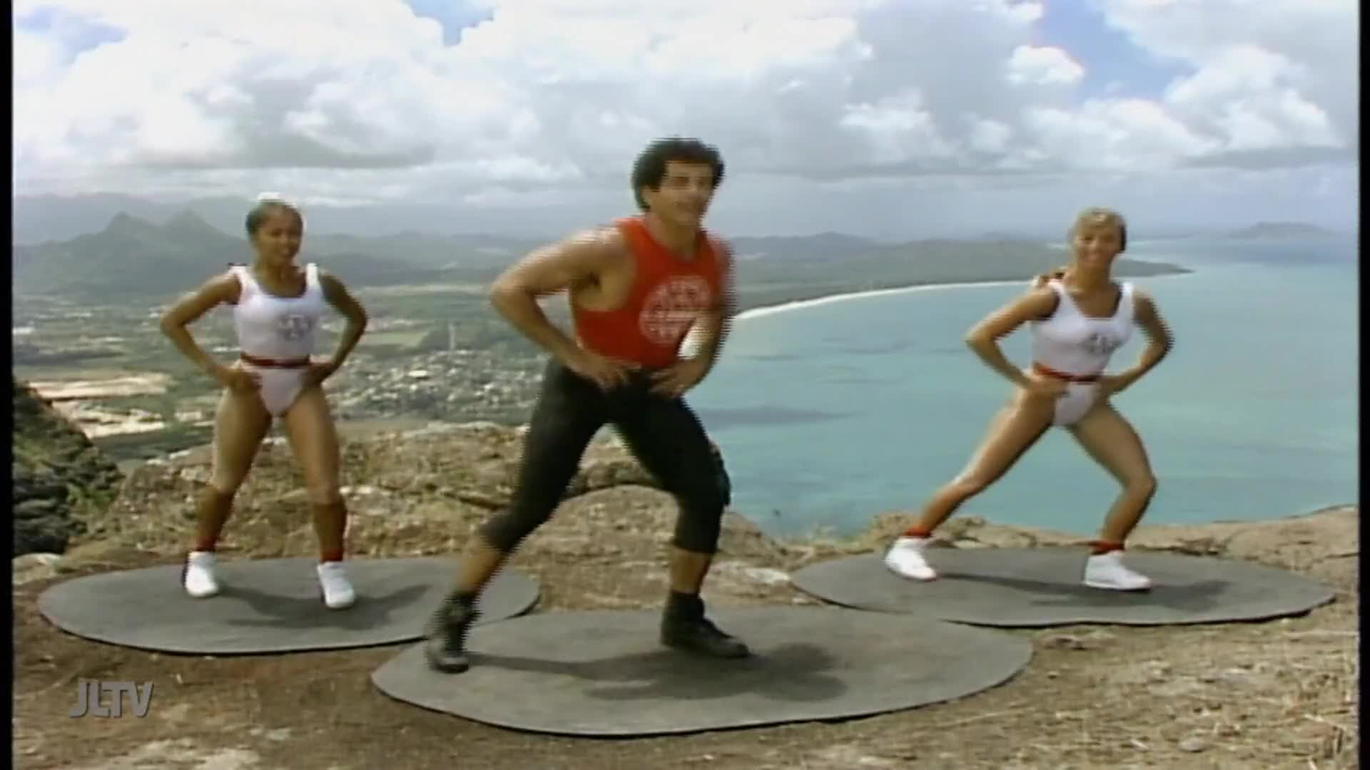 Three people in leotards and sneakers are stepping from side to side on mats on a mountain, with a beautiful ocean view behind them. This looks like a fitness show, maybe on Jewish Life Television.
