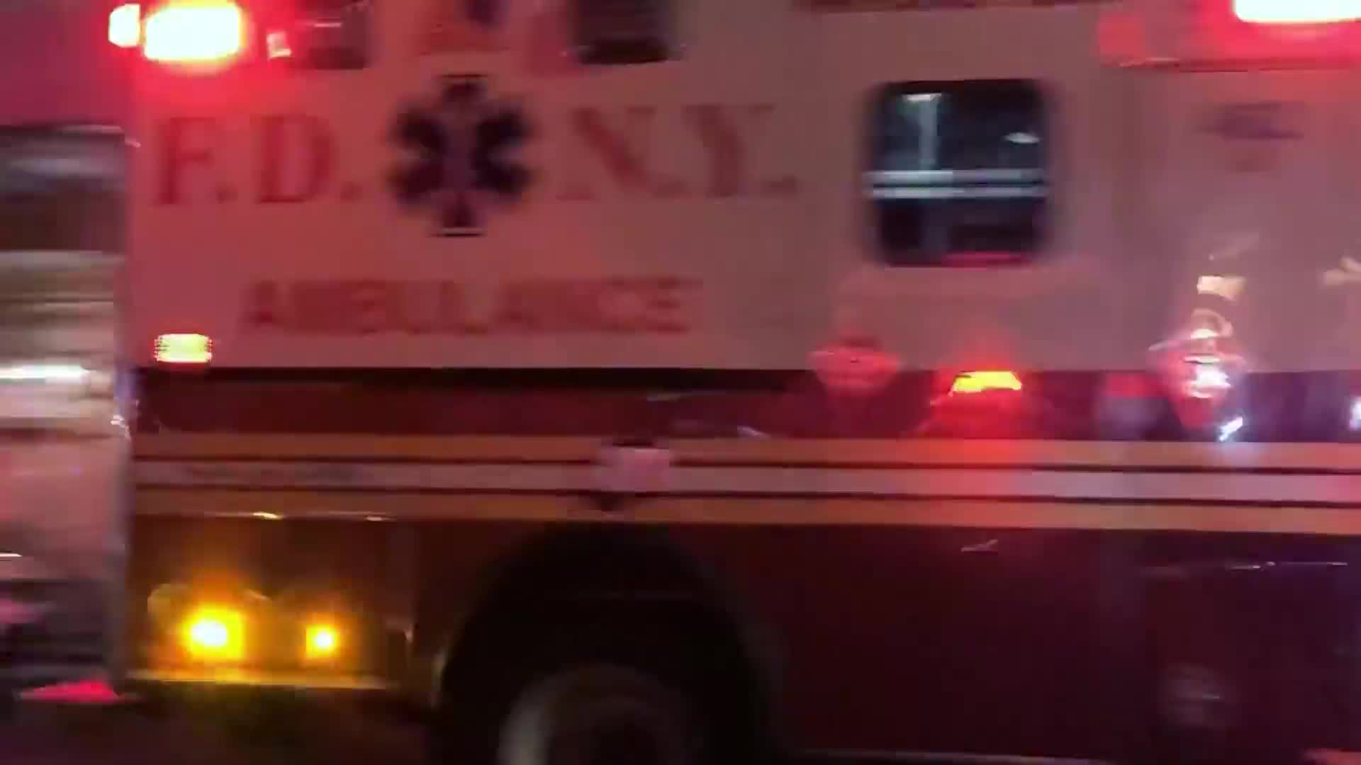 The side of an FDNY ambulance speeds by, its lights flashing. The vehicle's maroon and white paint is illuminated by the bright red and yellow emergency lights.
