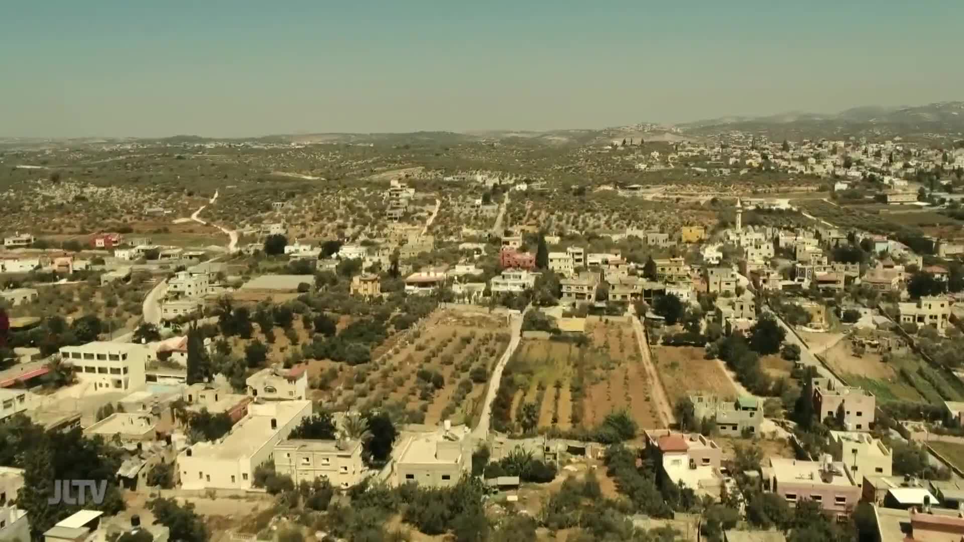 From this vantage, the camera surveys a sun-baked landscape dotted with buildings and olive groves. The scene, likely from a segment on Jewish Life Television, presents a clear view of a town nestled in the hills.
