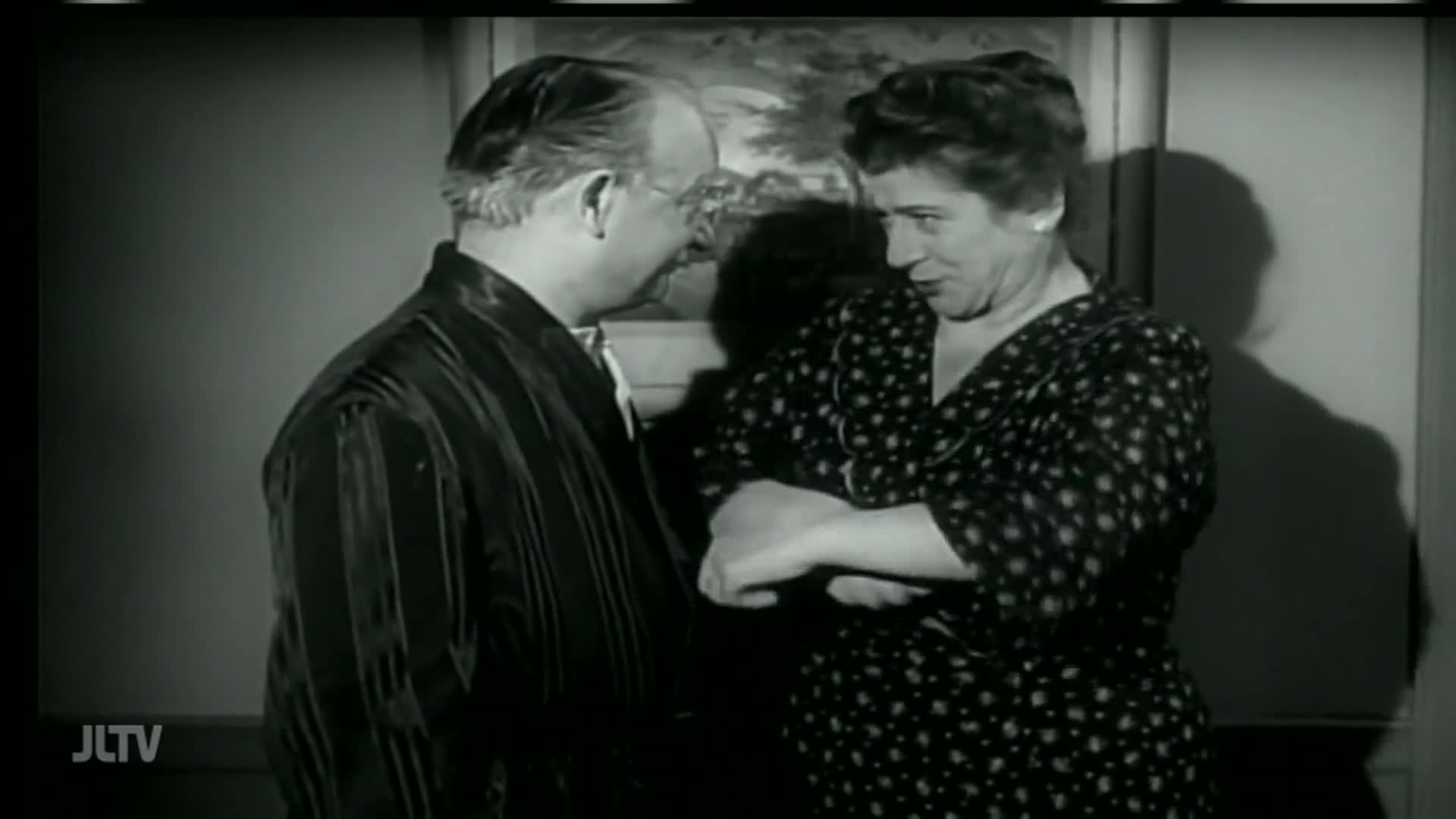On Jewish Life Television, a man in a silk robe and a woman in a polka-dot dress are in conversation, their faces close. The woman's expression is animated, and she gestures with her arms crossed.
