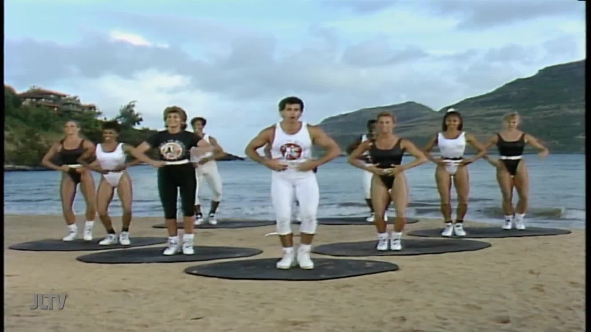 On a beach, a group of people in workout attire are doing exercises, guided by a man in a white tank top. The scene is being broadcast on Jewish Life Television.
