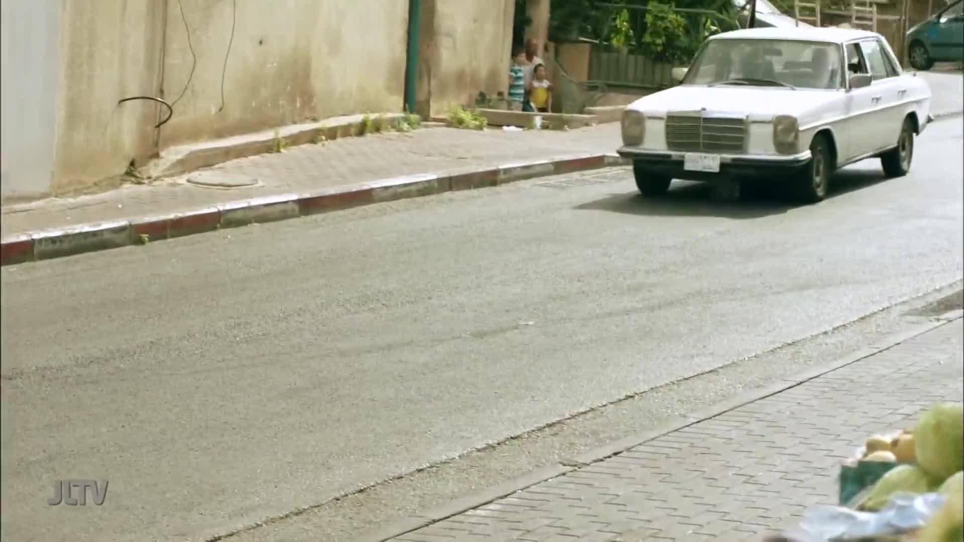 A white Mercedes glides down the street, its front license plate visible. Children stand on the sidewalk, watching the car pass by on what looks like a segment from Jewish Life Television.
