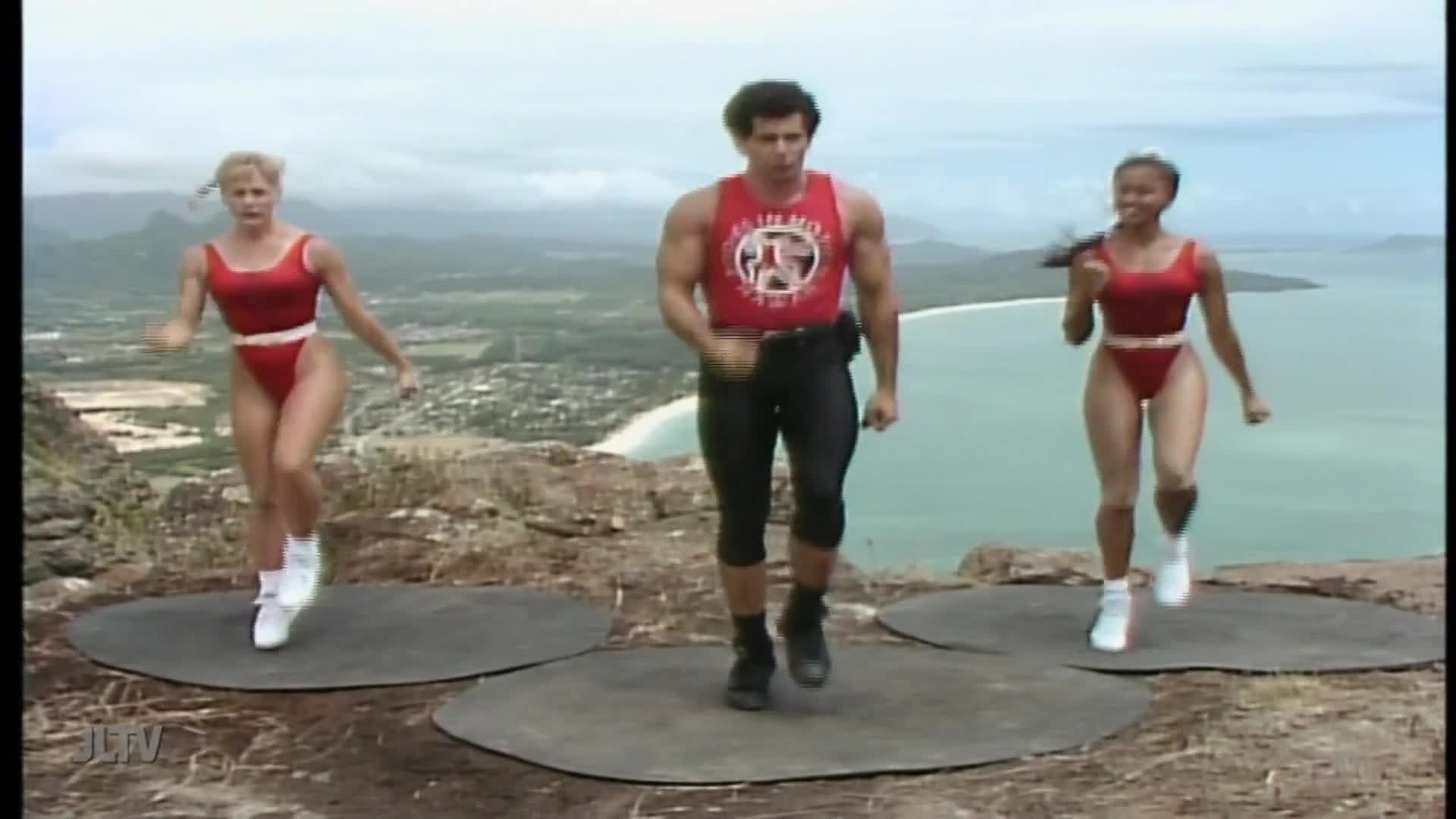 Three people in red leotards and black leggings are jogging on mats, with a coastal vista in the background. The man in the middle wears a tank top with a logo, while the women bounce in unison.
