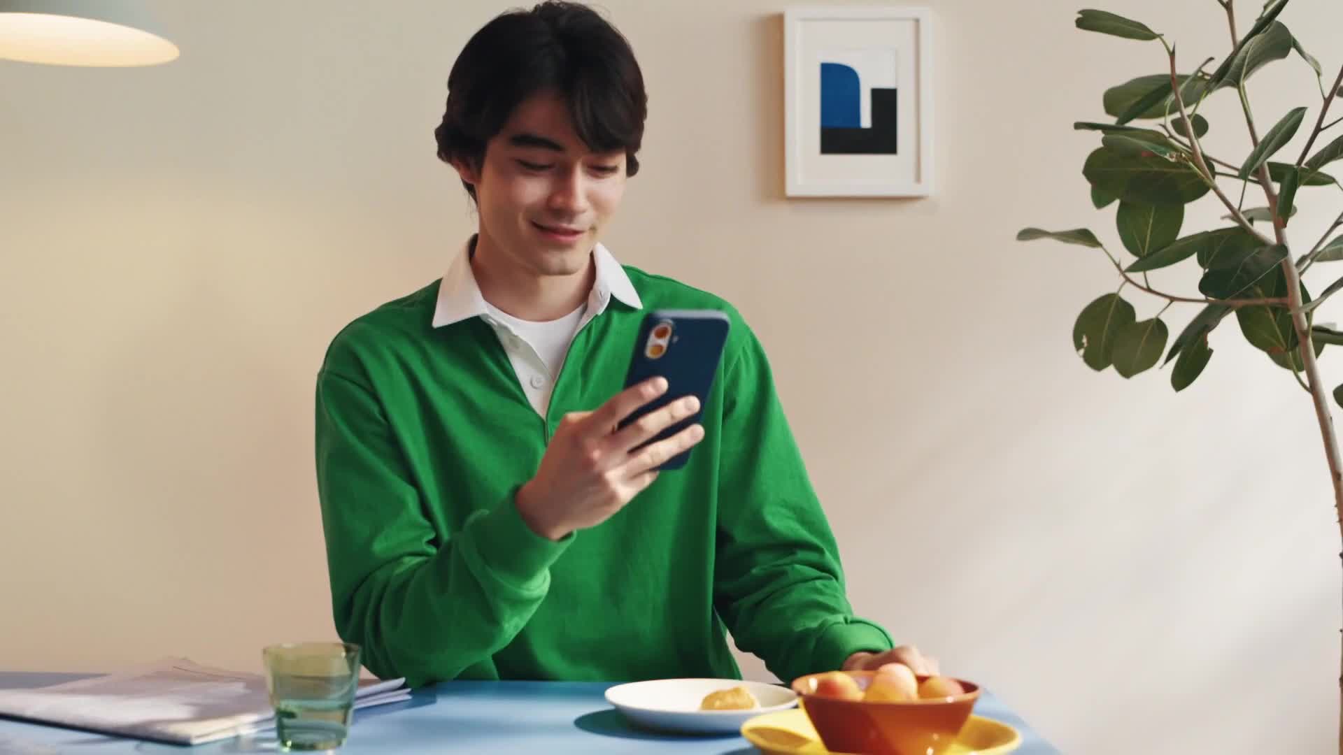 A man in a green shirt is seated at a table, smiling as he looks at his phone. A small bowl of fruit and a plate with a pastry sit nearby.
