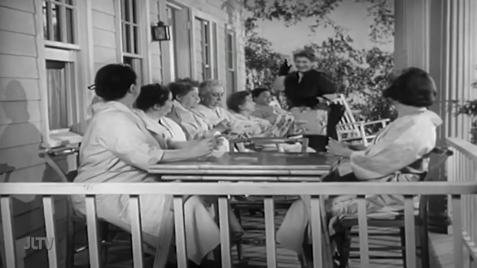 On a porch, a group of women in matching robes sit around a table, some holding cards. A woman in a dark dress and apron approaches, possibly offering something.
