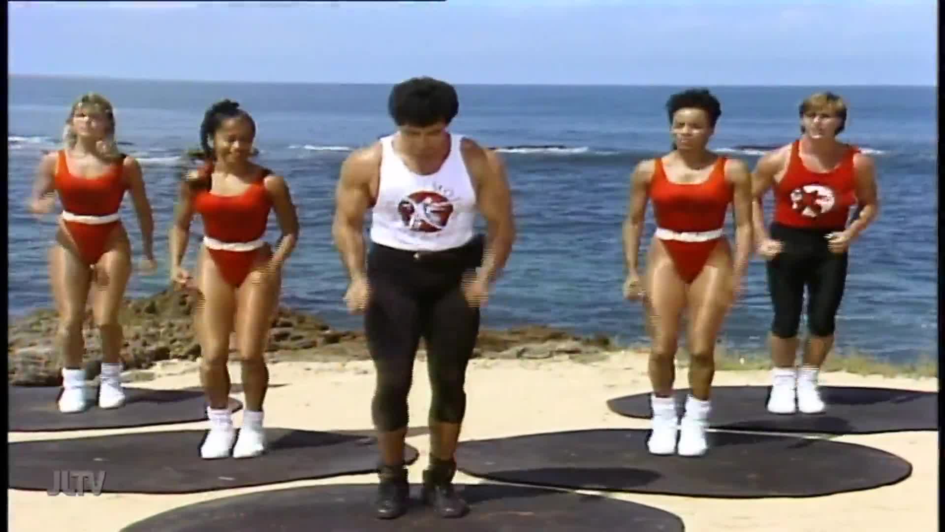 On a beach, a group of people in brightly colored workout gear are jogging in place on mats. The man in the center, wearing a tank top, is leading the exercise routine.
