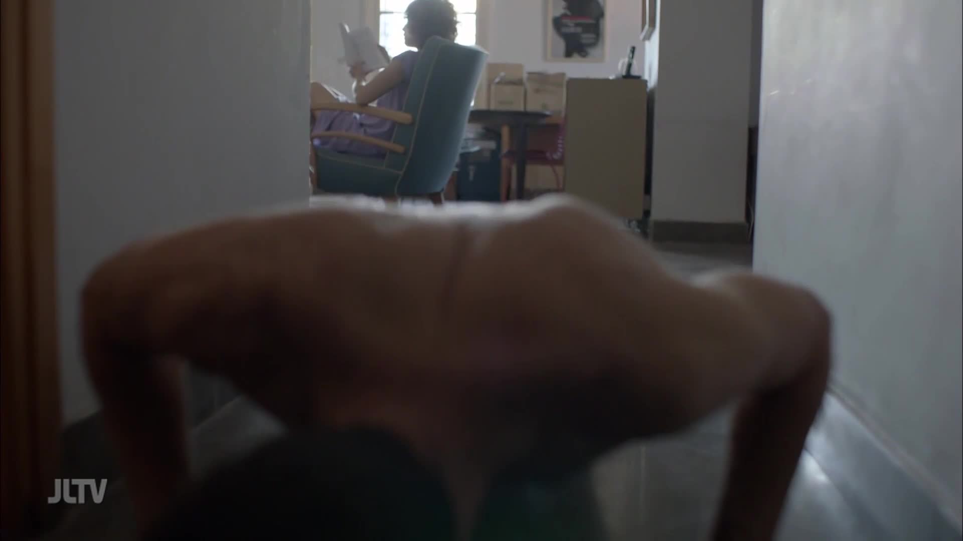 A person does push-ups in a hallway, their back and arms taking up most of the frame. In the background, a woman sits in a chair, reading a book.
