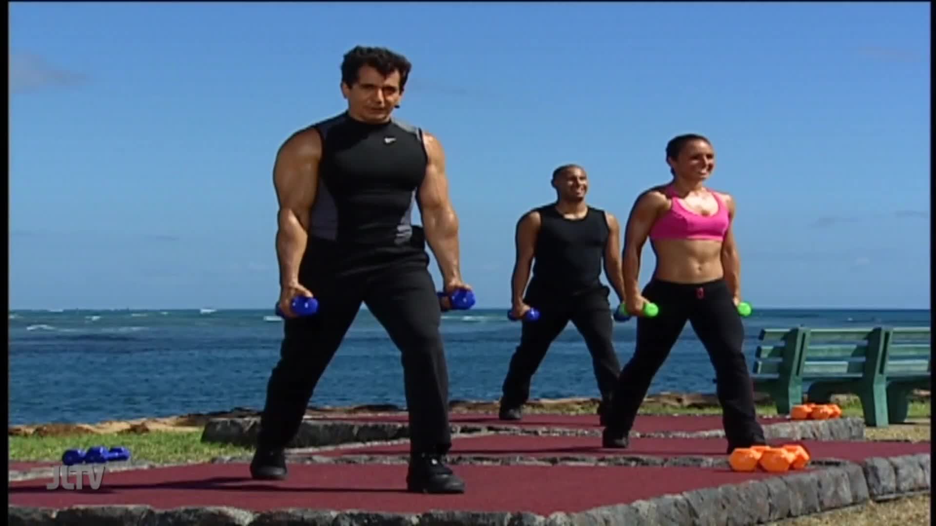 On a sunny day, three people are exercising with dumbbells on a raised platform near the ocean. The man in the center, leading the workout, is on Jewish Life Television.
