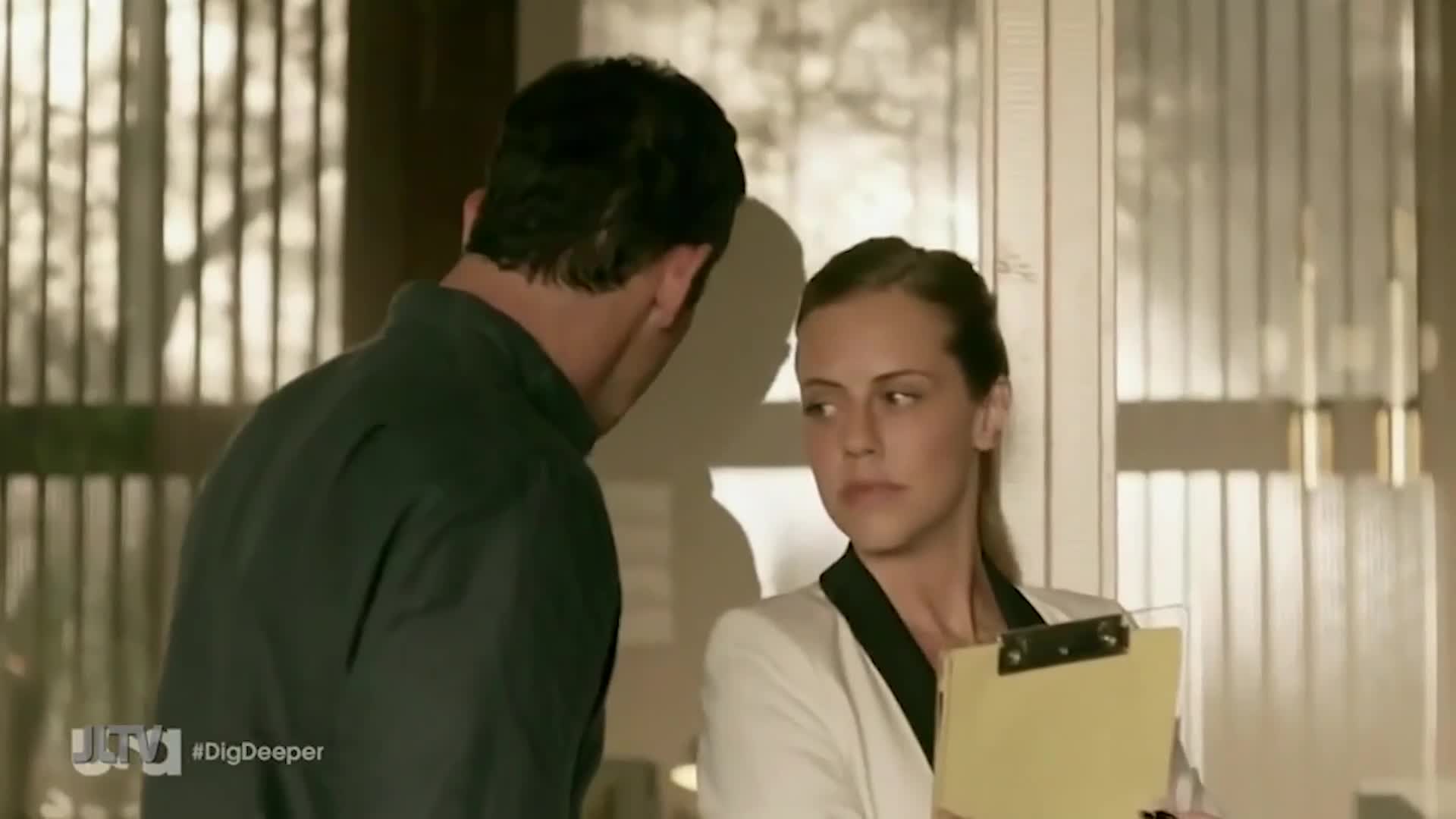 A man in a dark shirt leans toward a woman, who holds a clipboard and looks at him with a serious expression. The scene seems to be from a United States Jewish Life Television show, "Dig Deeper."
