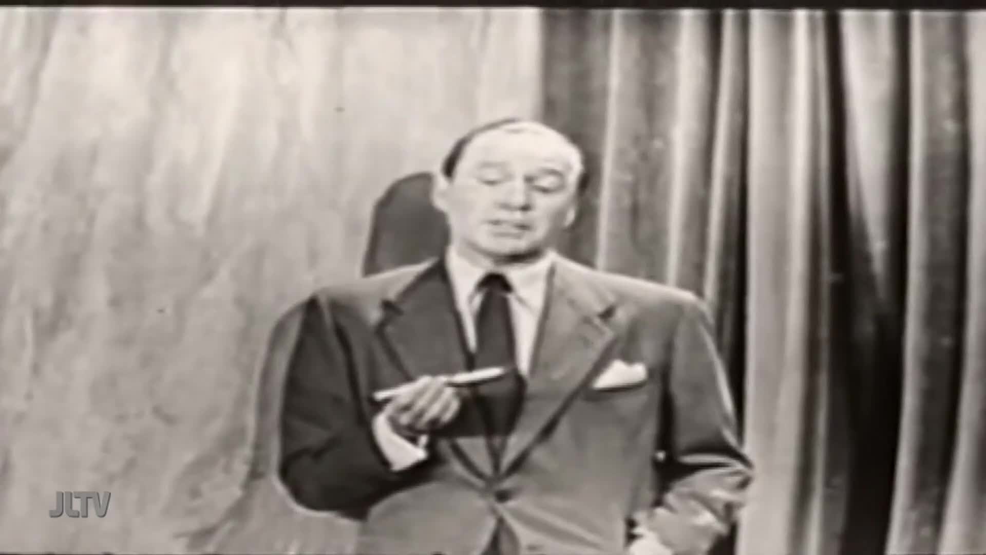 A man in a suit, holding a pen, gestures with his right hand. He's speaking on a set, with a painted backdrop and curtains behind him, likely on Jewish Life Television.
