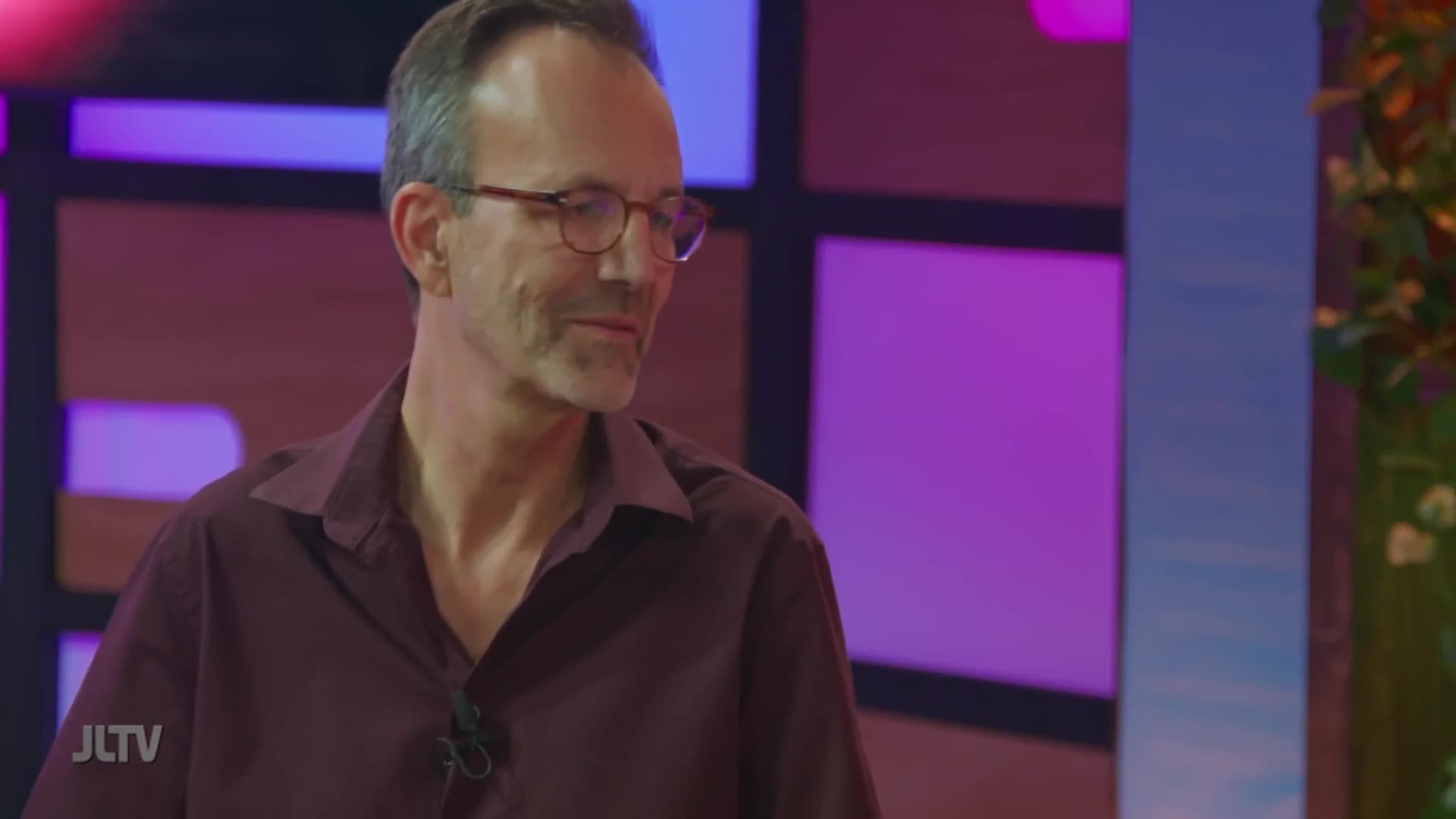 A man in glasses and a maroon shirt is looking off to the side, a small microphone clipped to his collar. The set behind him is lit with purples and pinks, and the logo of Jewish Life Television is visible.
