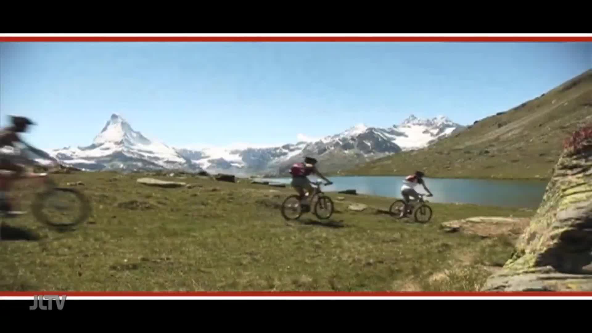 Mountain bikers are pedaling across a grassy field, heading towards a lake with snow-capped mountains in the background. The scene is framed by the logo of Jewish Life Television.

