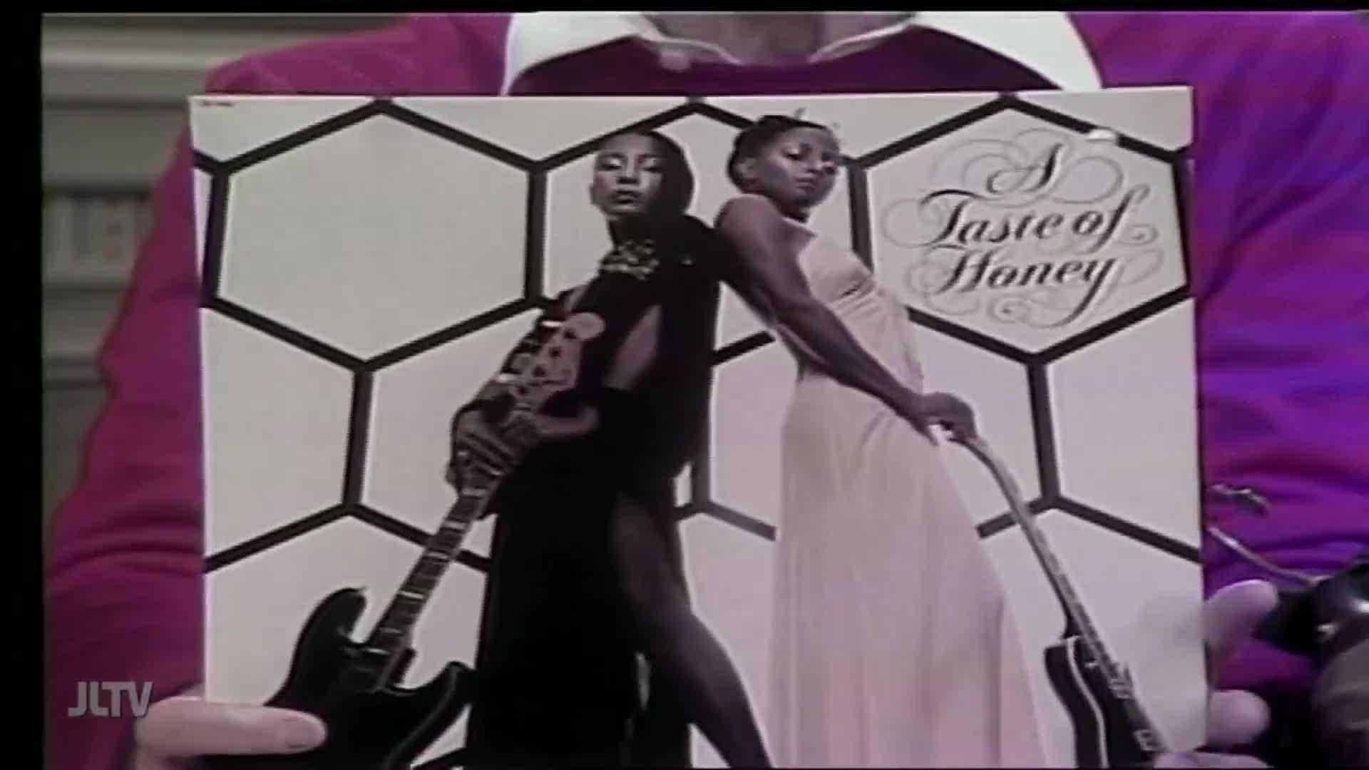 A man in a pink sweater holds up an album cover for "A Taste of Honey" on Jewish Life Television. The cover art features two women, each holding a guitar, standing back-to-back.
