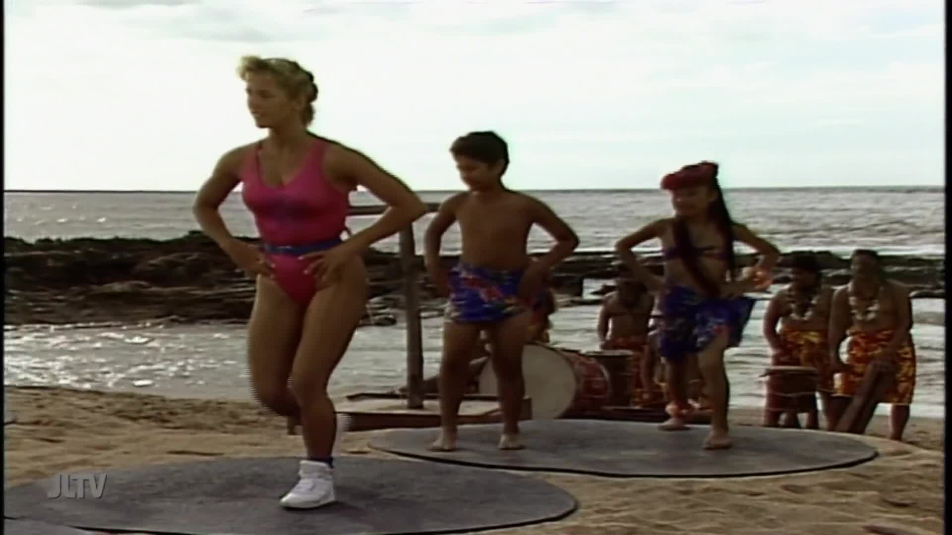 On a beach, a woman in a pink leotard and white sneakers leads a dance. A boy and a girl, along with other dancers, follow her moves on circular platforms as drummers provide the beat.
