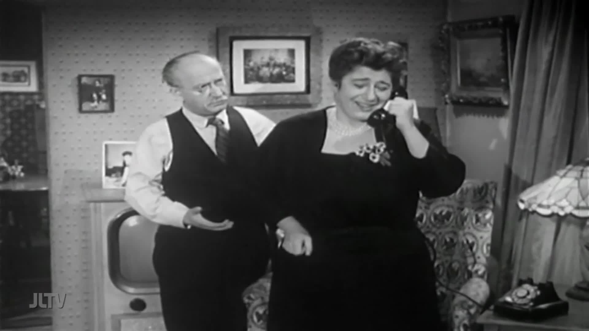 A woman in a dark dress holds a phone to her ear, her face a mix of emotions. Beside her, a man in a vest looks on, his hand gesturing as if he's about to say something.
