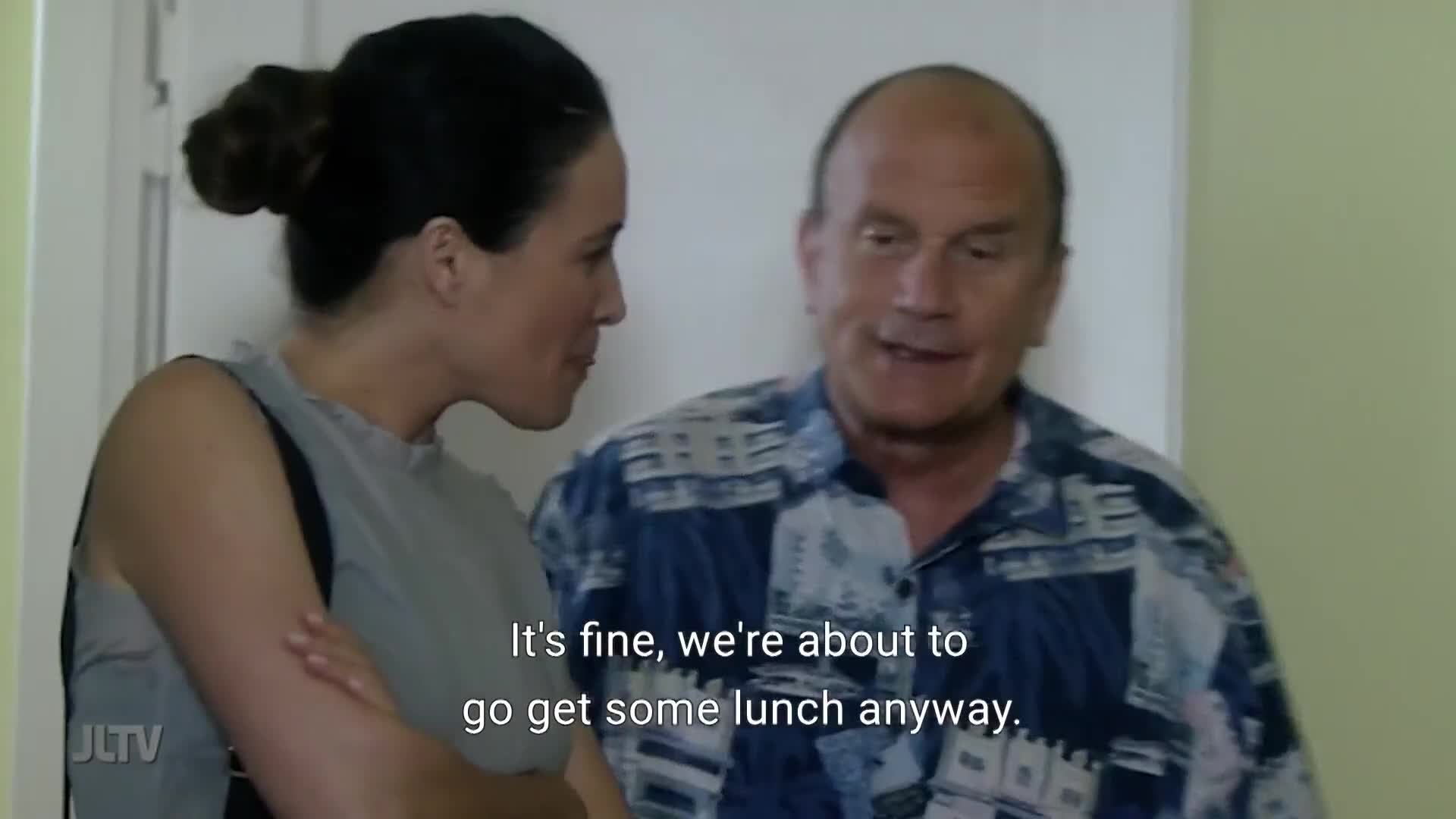 A man in a patterned shirt is speaking, gesturing with his hands. Beside him, a woman with her arms crossed listens intently, her expression calm.
