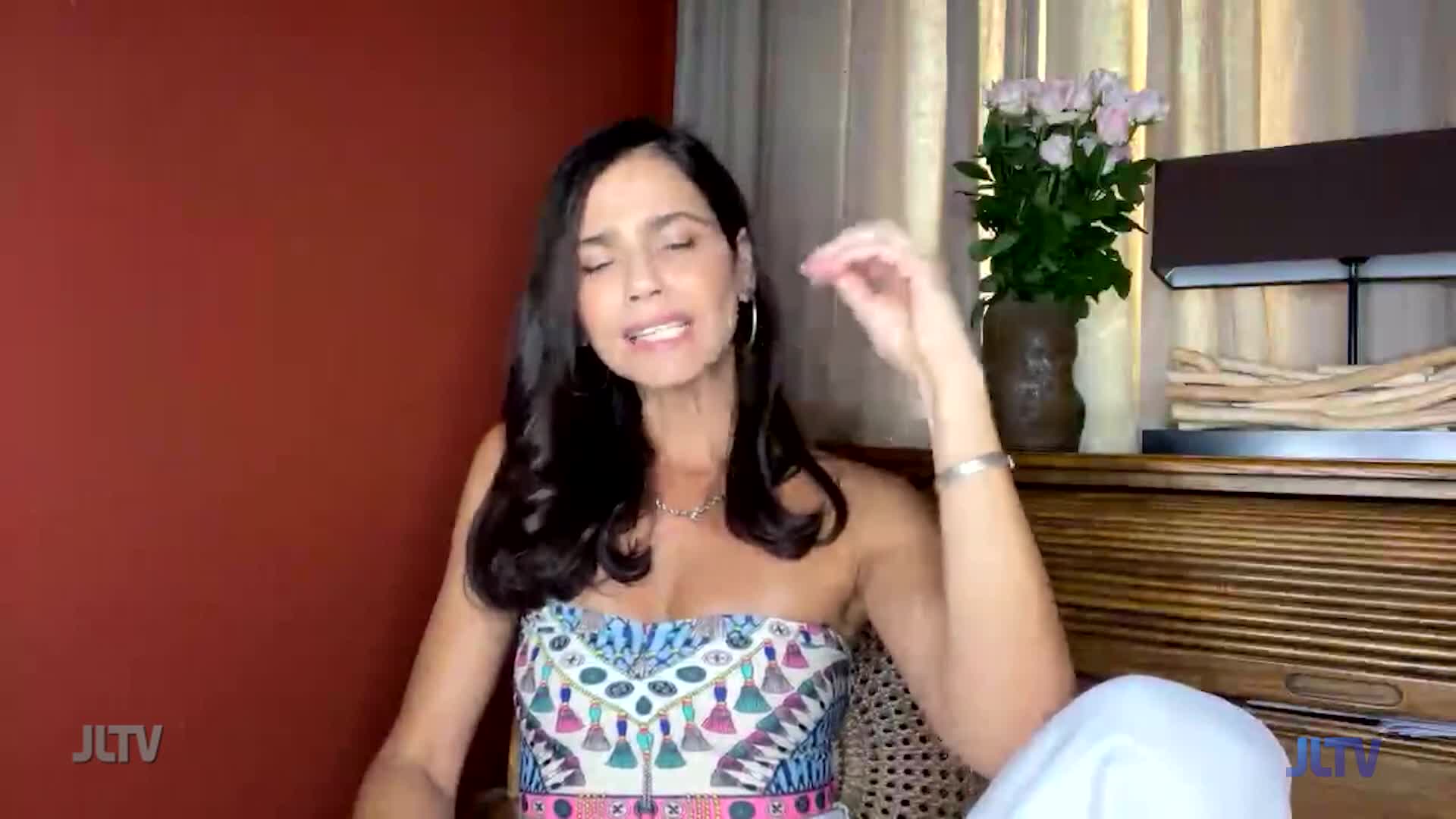 A woman on Jewish Life Television gestures with her right hand, her expression animated as she speaks. Behind her, a vase of pink flowers sits atop a dark wooden surface.
