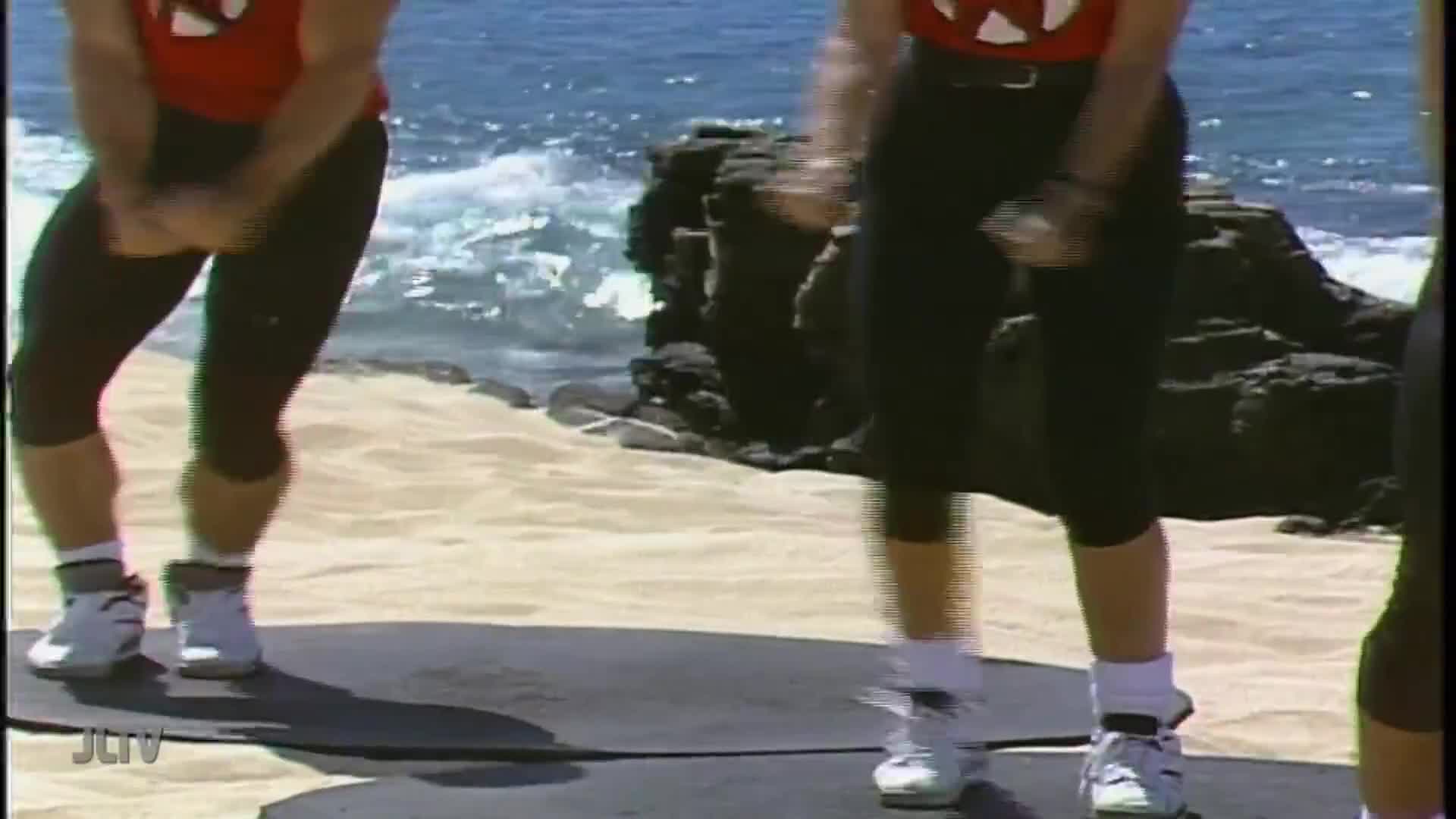 On a beach, people in matching outfits are bouncing on mats. The ocean waves crash in the background as they lift their knees and pump their arms.
