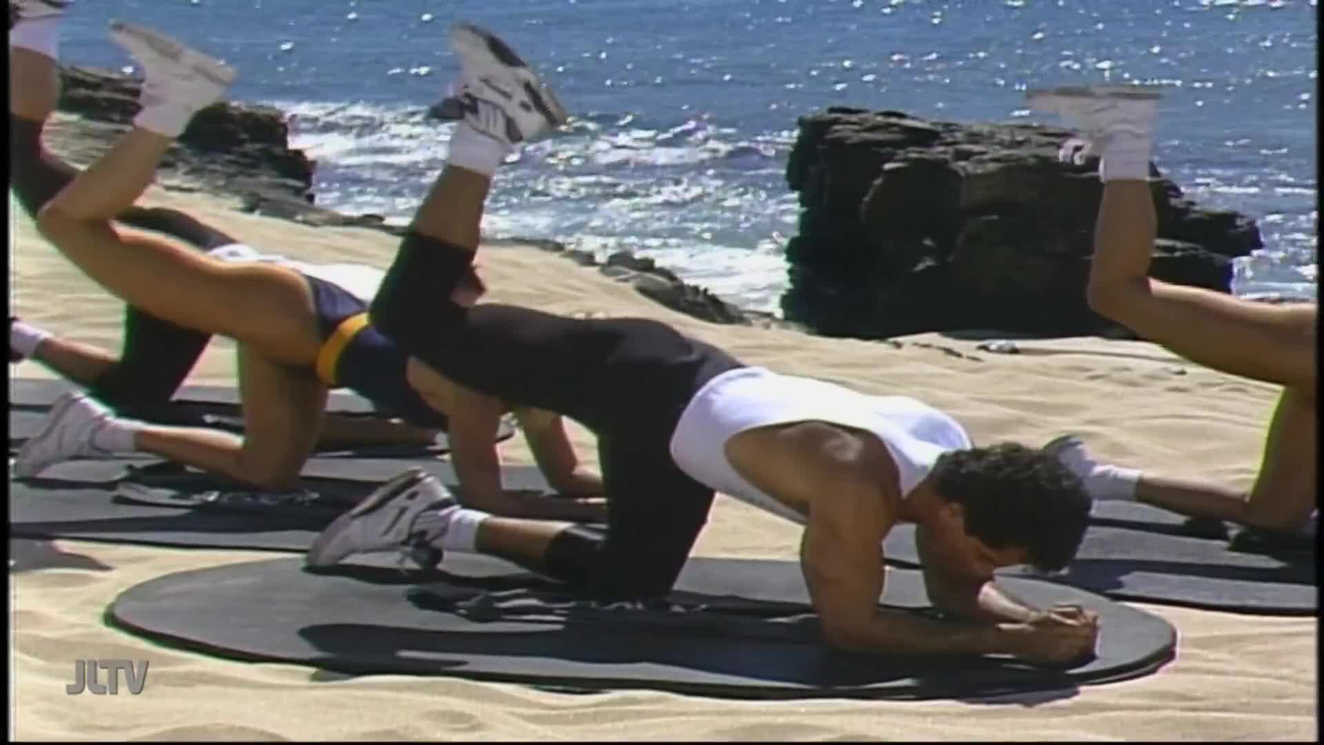 On a beach, people are on their hands and knees, lifting their legs in a workout. The scene is being filmed for Jewish Life Television, with the ocean shimmering in the background.
