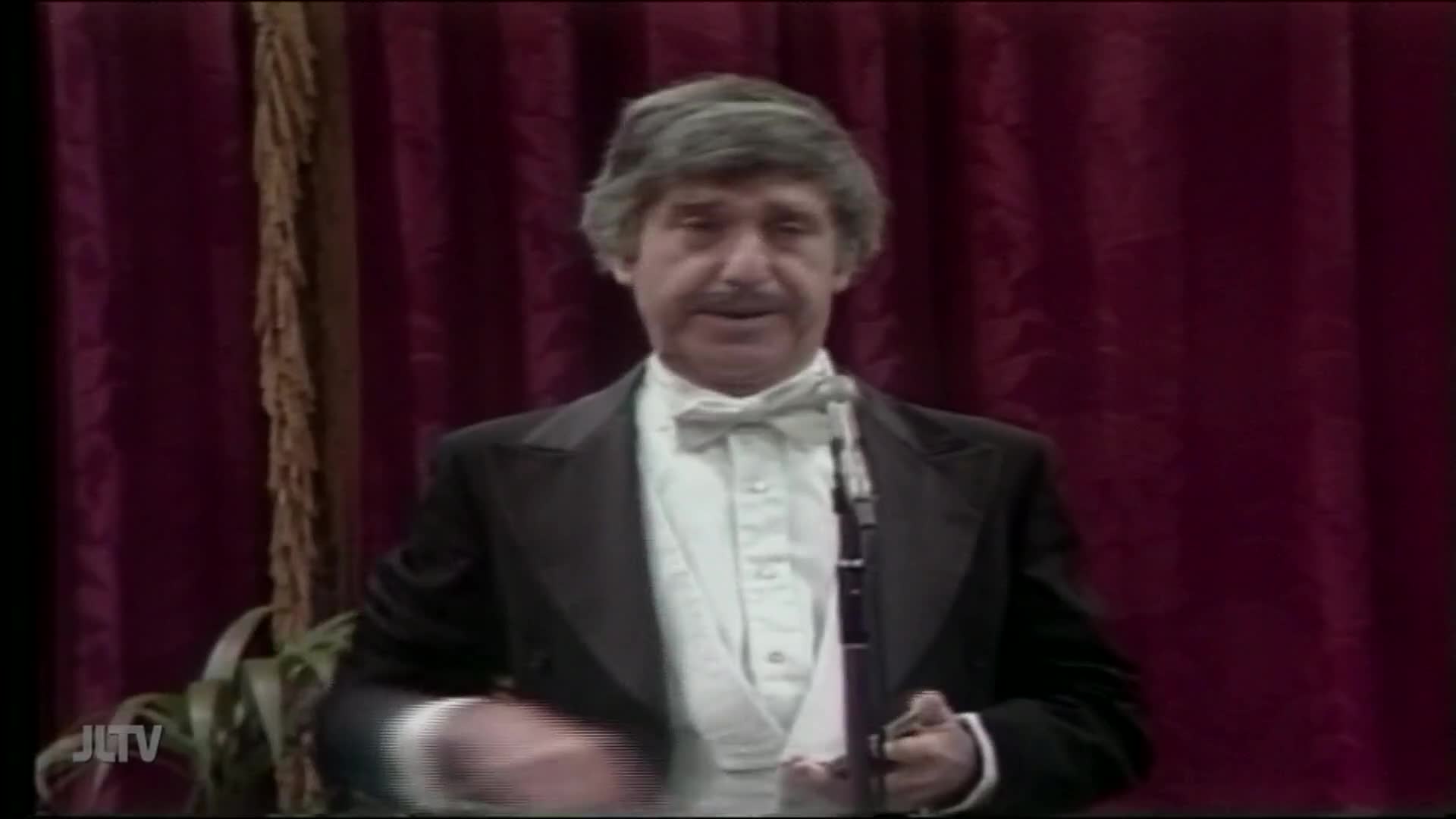 A man in a tuxedo stands before a microphone, speaking on Jewish Life Television. He's holding the microphone with one hand and gesturing with the other, against a backdrop of red curtains.
