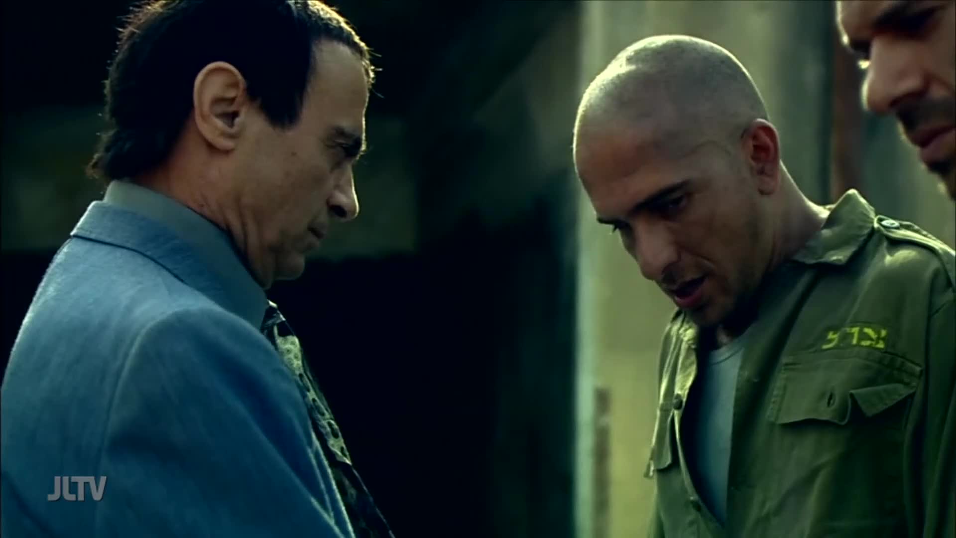 A man in a blue suit looks down, listening intently to the man in the green military-style shirt. The scene, from Jewish Life Television, is heavy with unspoken tension.
