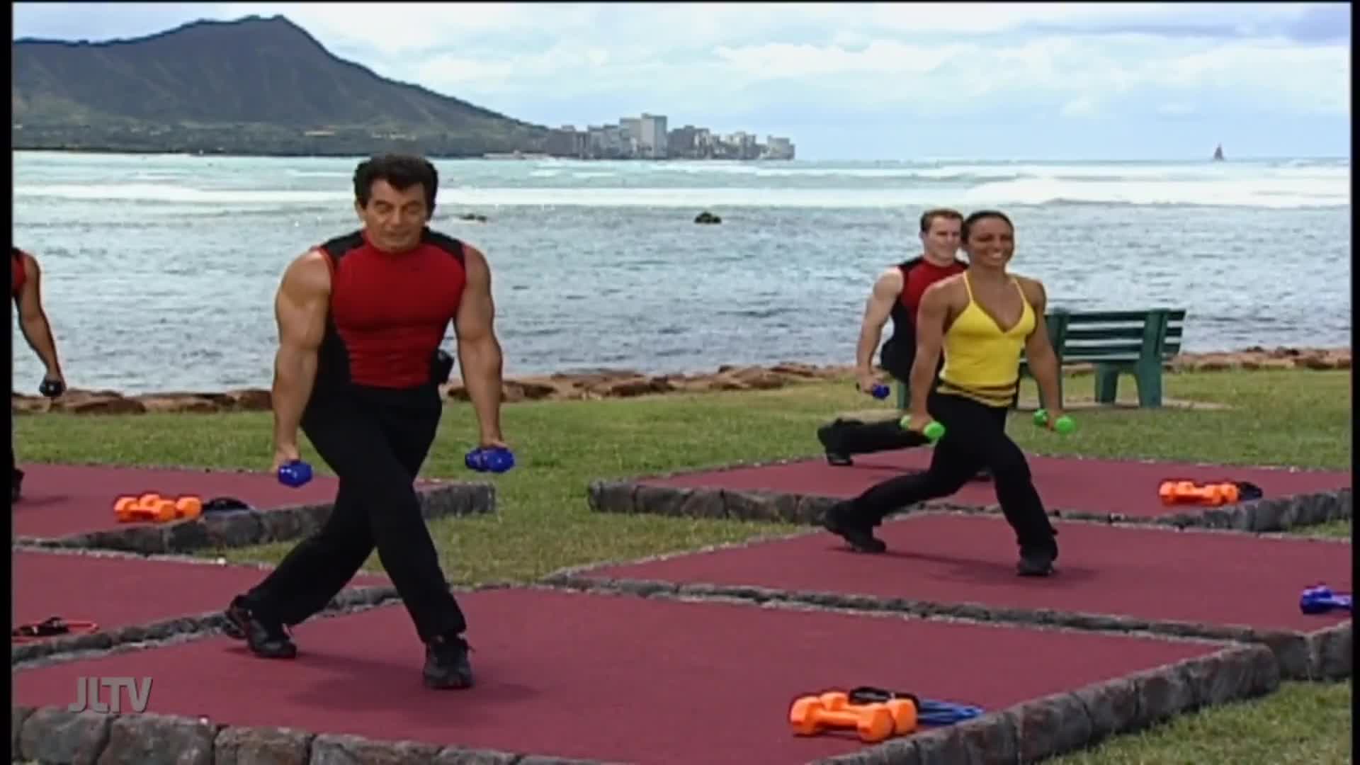 A man in a red shirt and black pants steps forward, holding blue dumbbells, as he lunges. Other people, also exercising with weights, are visible on the red mats, with the ocean and a mountain in the background.
