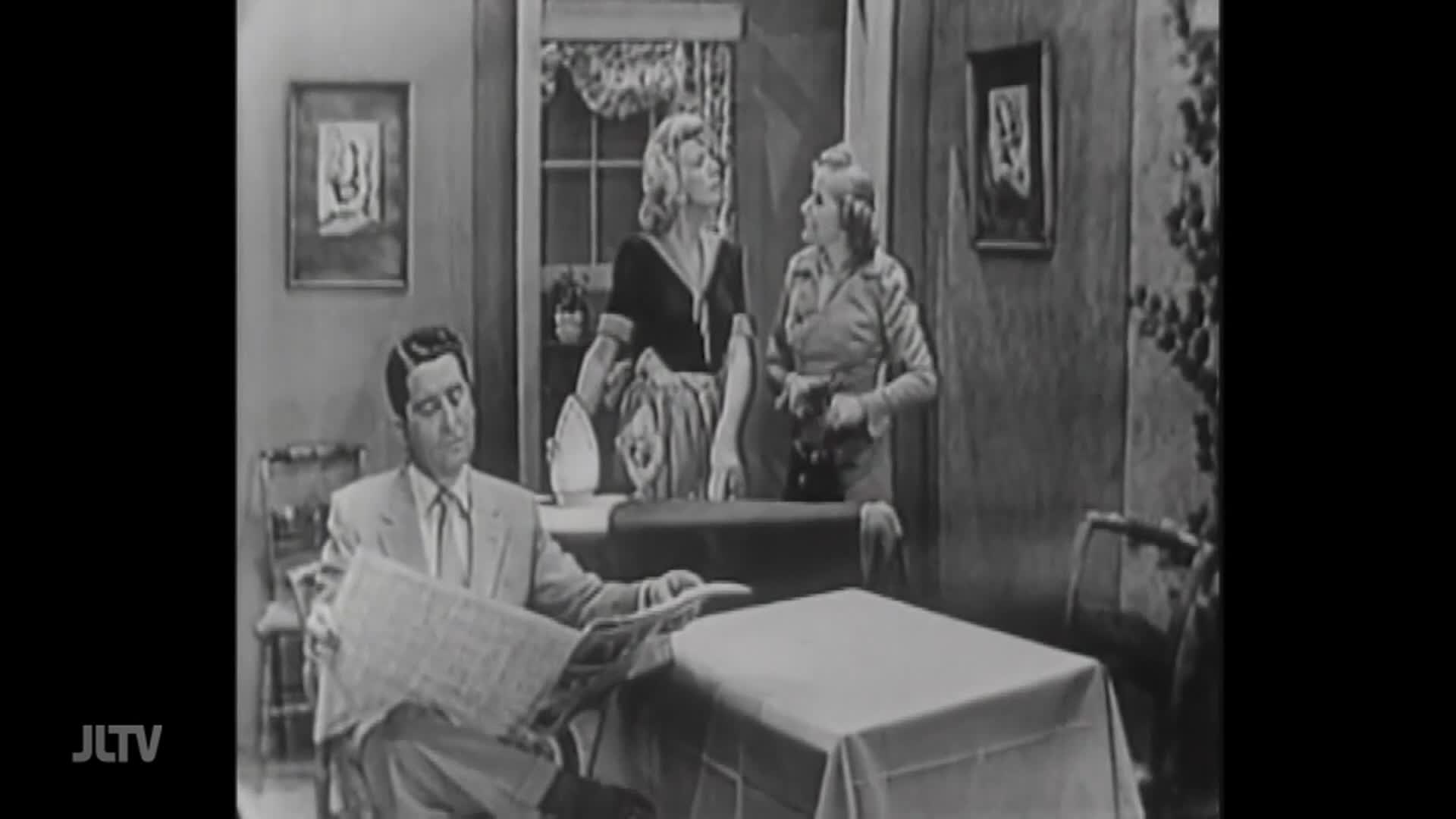 A man in a suit sits at a table, reading a newspaper. Two women stand behind a table, one holding an iron, the other looking at a camera.
