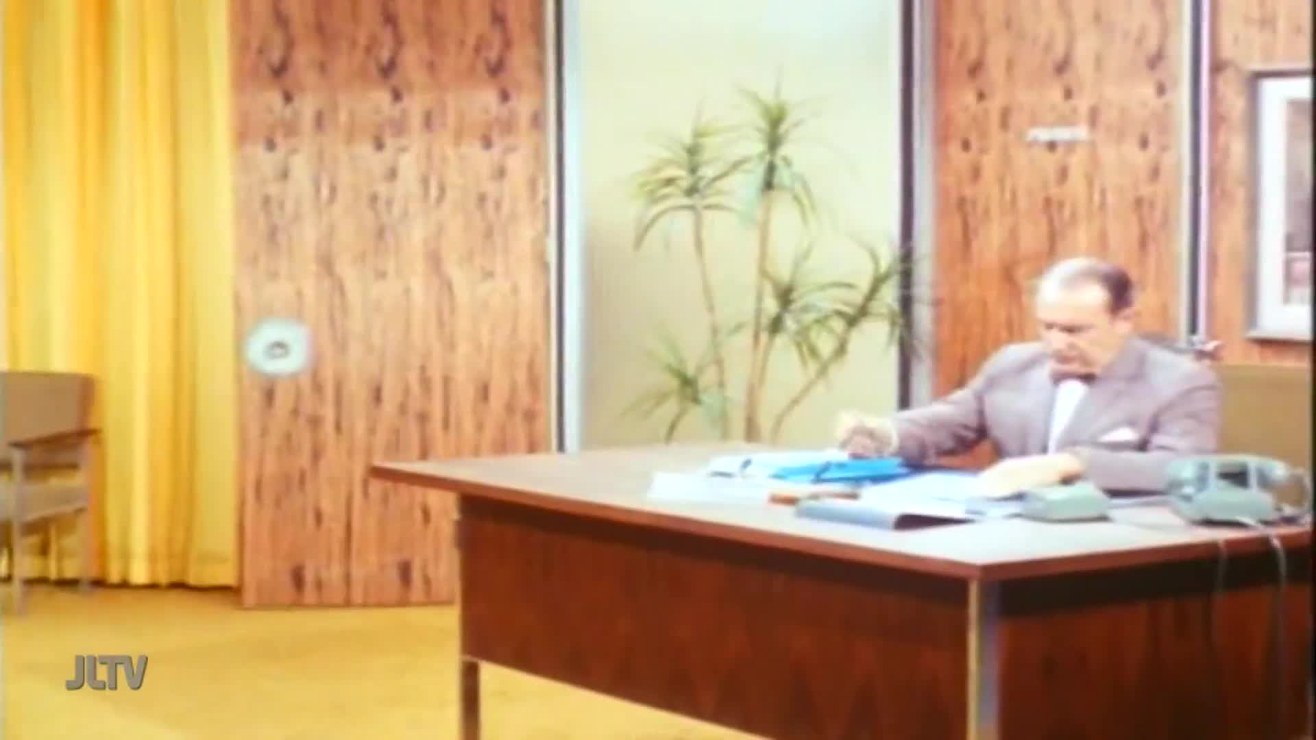 A man in a suit sits at a large desk, carefully sorting through papers. The scene, likely from a United States-based Jewish Life Television program, is set in a wood-paneled office.
