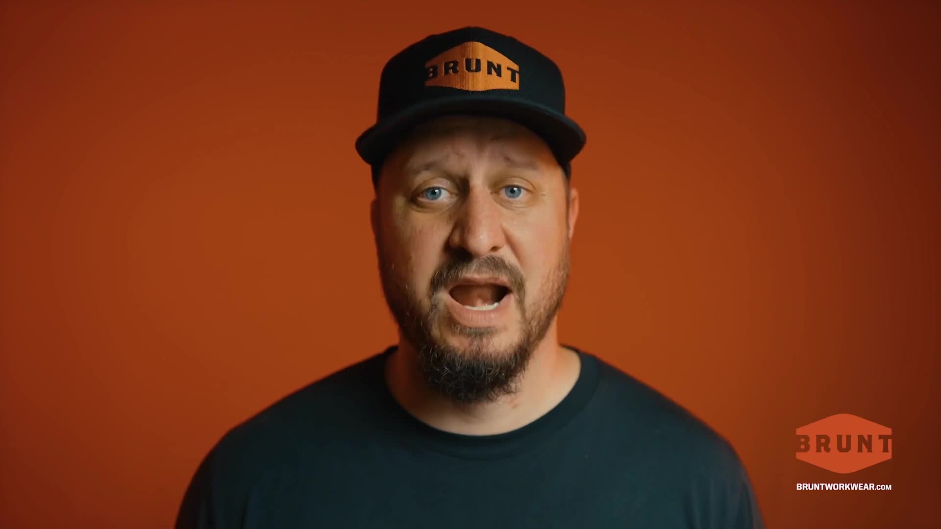 A man with a beard and a baseball cap is speaking directly to the camera against a solid orange background. His mouth is open, and he seems to be mid-sentence.
