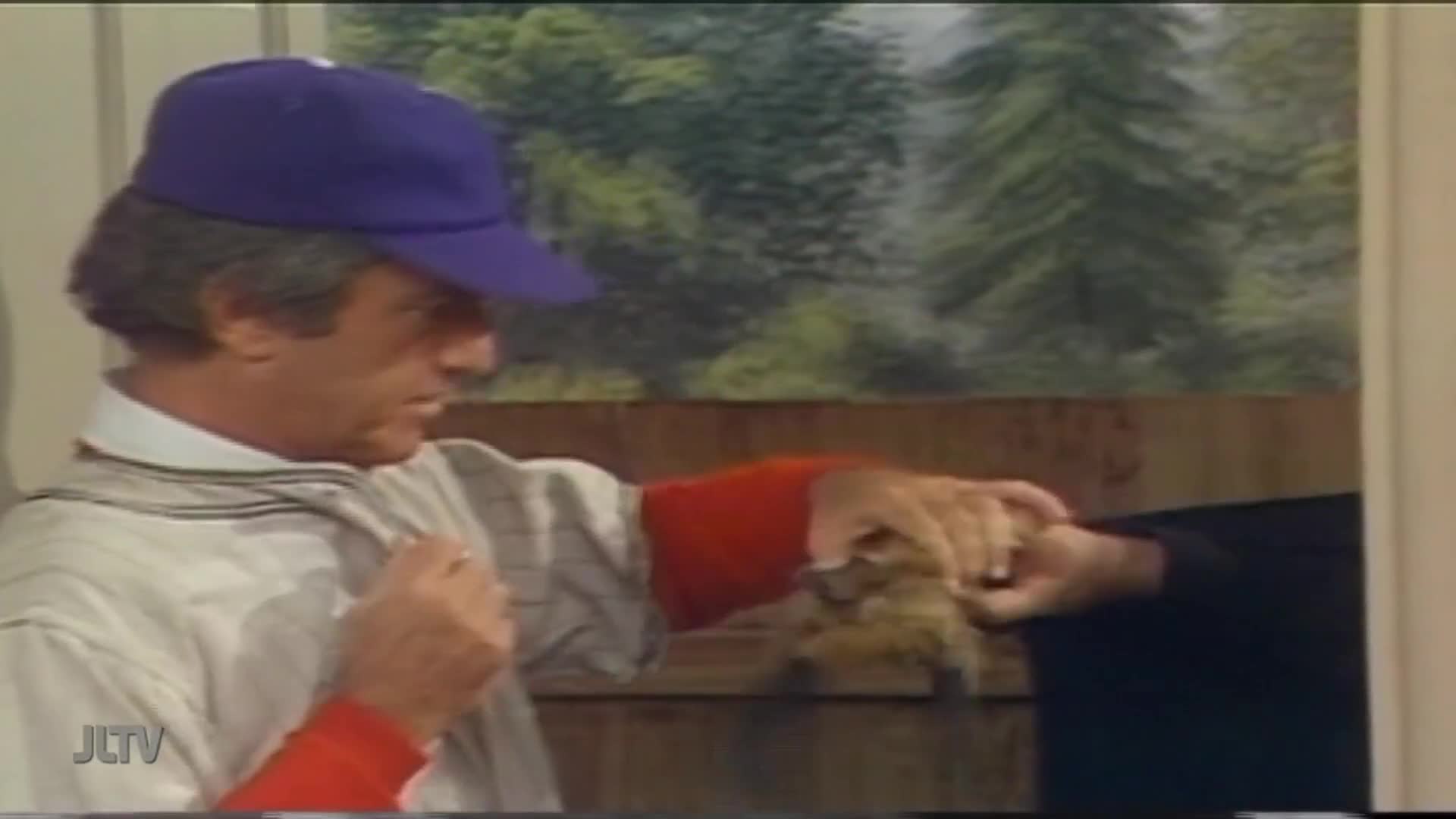 A man in a baseball jersey and cap is holding something small and furry, possibly a pet, with another person. The scene is from a Jewish Life Television program.
