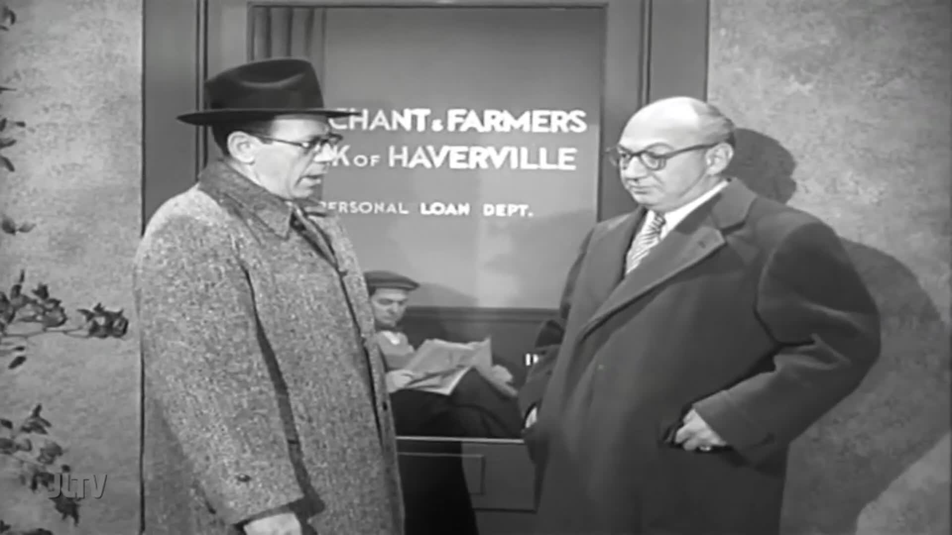 Two men in coats and fedoras stand outside the "Chant & Farmers Bank of Haverville" Personal Loan Dept. One speaks animatedly while the other listens, his hand in his pocket.
