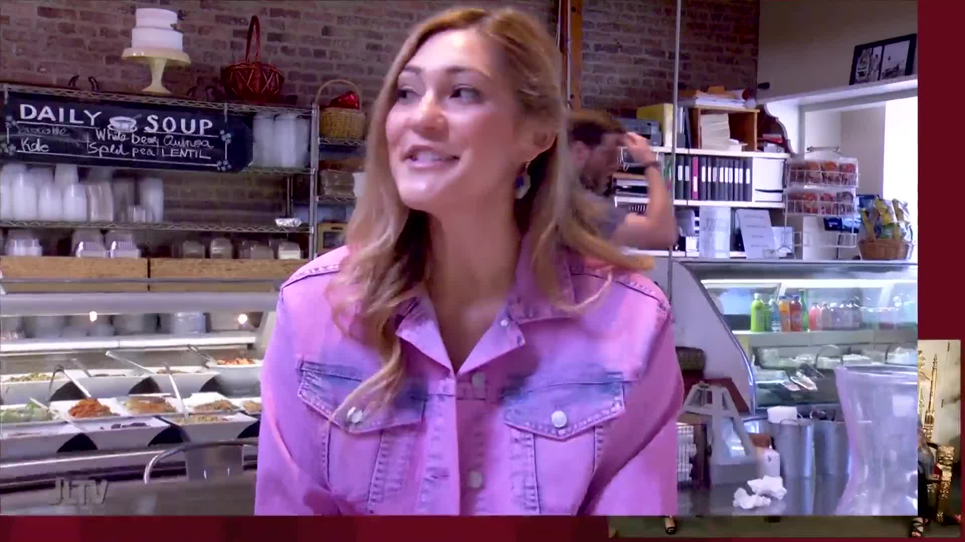 A woman in a pink denim jacket is speaking, likely for Jewish Life Television. Behind her, a deli counter displays various prepared foods.
