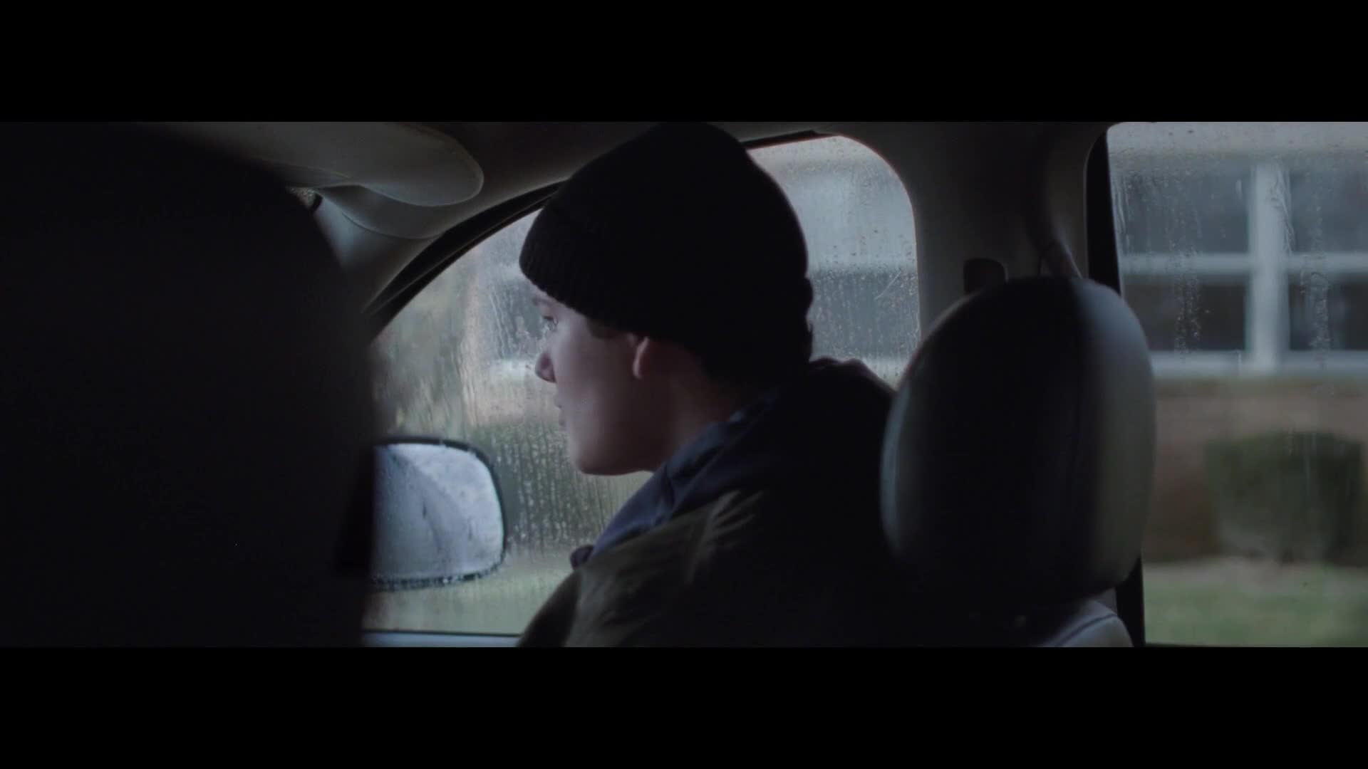 A young man in a black beanie stares out the rain-streaked window. The car's interior is dim, but the rain outside makes the world visible.
