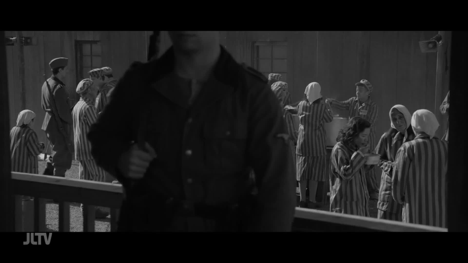 Prisoners in striped uniforms and headscarves are receiving a meal from a woman in a similar outfit. A man in a dark uniform walks past, his back to the camera.
