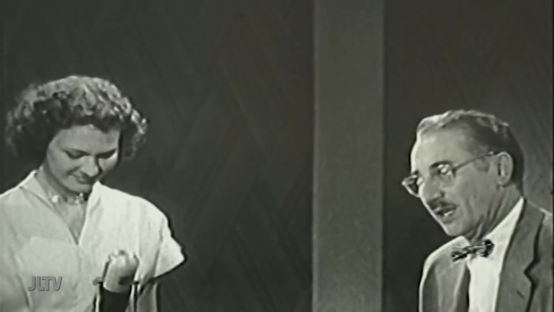 A woman in a light-colored dress holds a microphone, looking down with a slight smile. Across from her, a man in glasses and a bow tie speaks animatedly.
