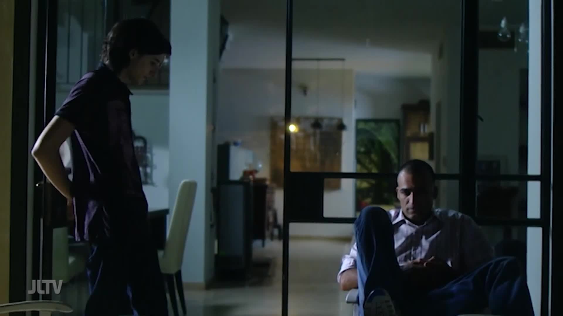 A man in a button-down shirt sits in a chair, looking down, while another person stands in the doorway with their hands on their hips. The scene, framed by a sliding glass door, looks like a moment of quiet contemplation.
