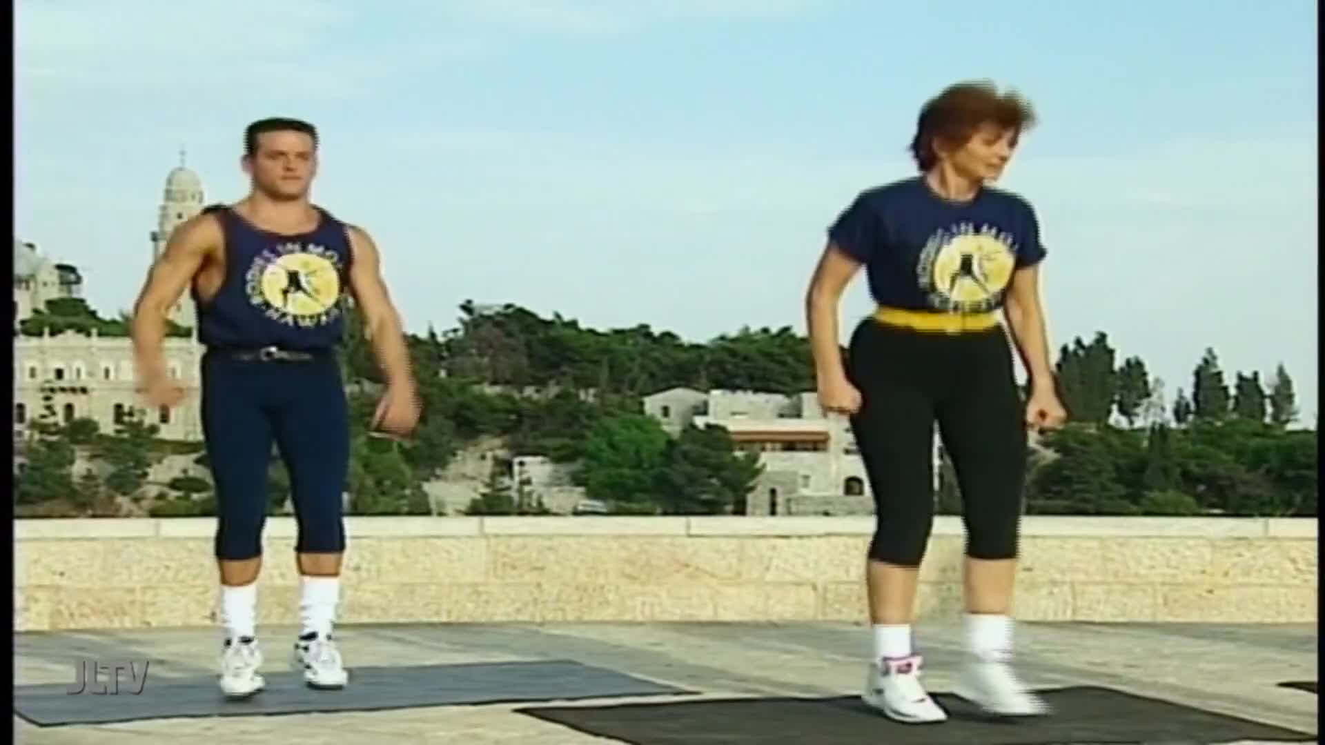 A man and woman, both in matching outfits, are bouncing on the spot. They're exercising outside, with a city and a tall building visible in the background.
