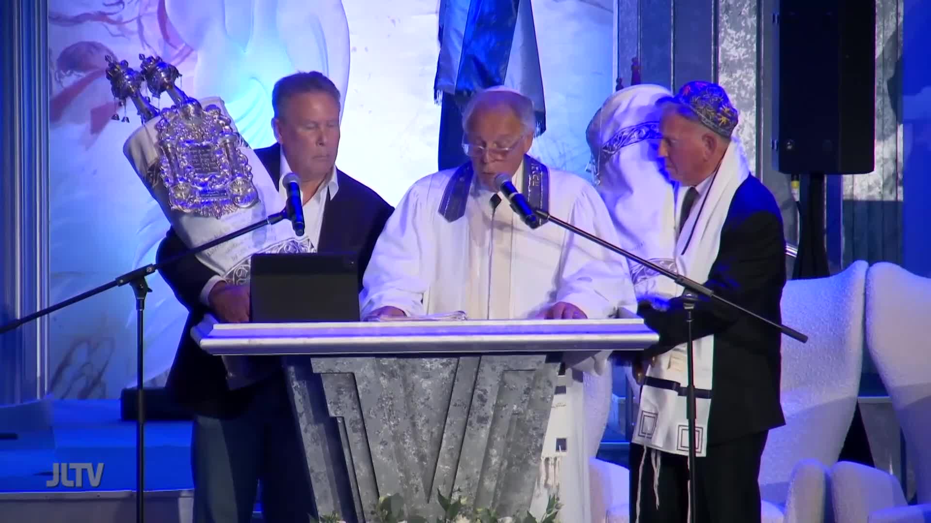 A rabbi in a white robe reads from a book behind a podium, while two men stand beside him, one holding a Torah scroll. The scene is being broadcast on Jewish Life Television, with a backdrop of blue and white.
